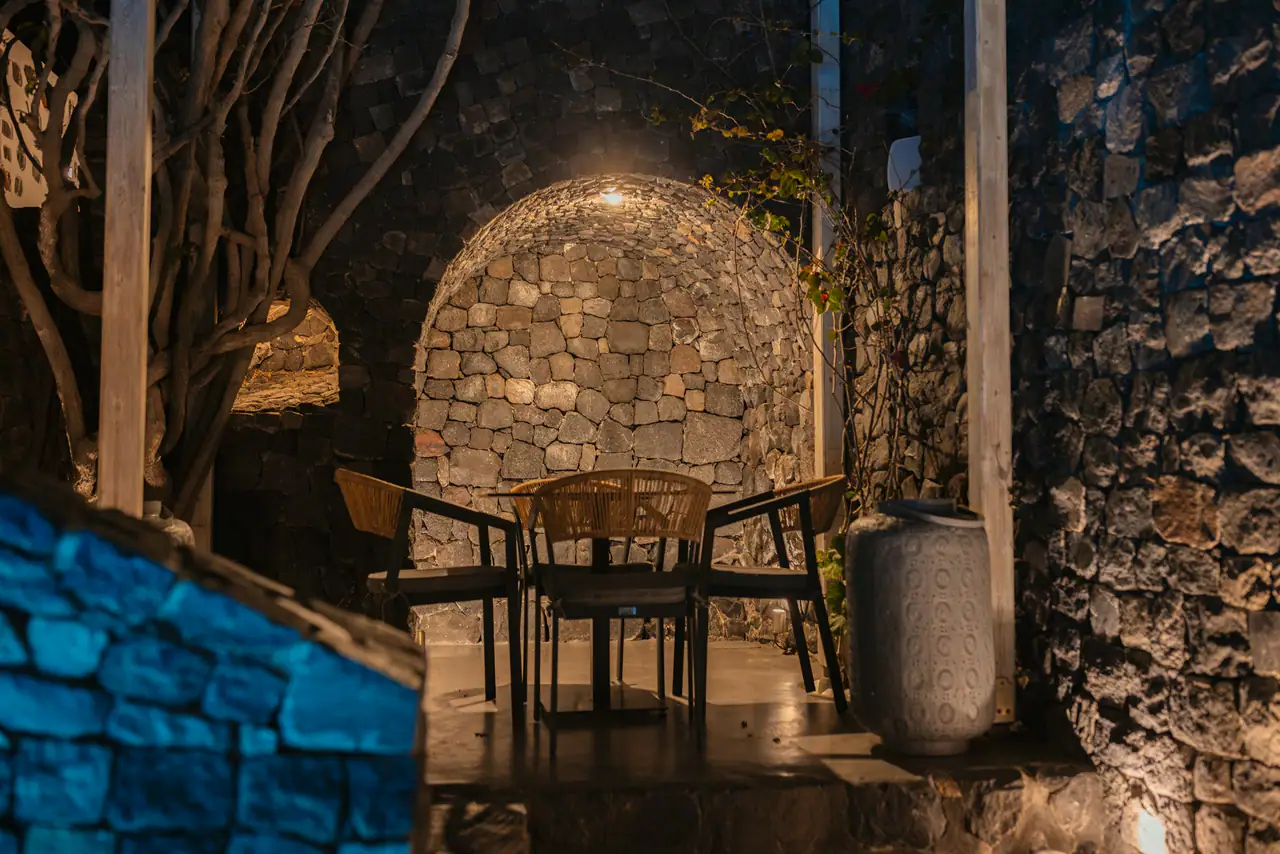 A candlelit stone archway at night with a small wooden table and chairs, warm lamplight on dark volcanic stonework, in a Mediterranean-style courtyard