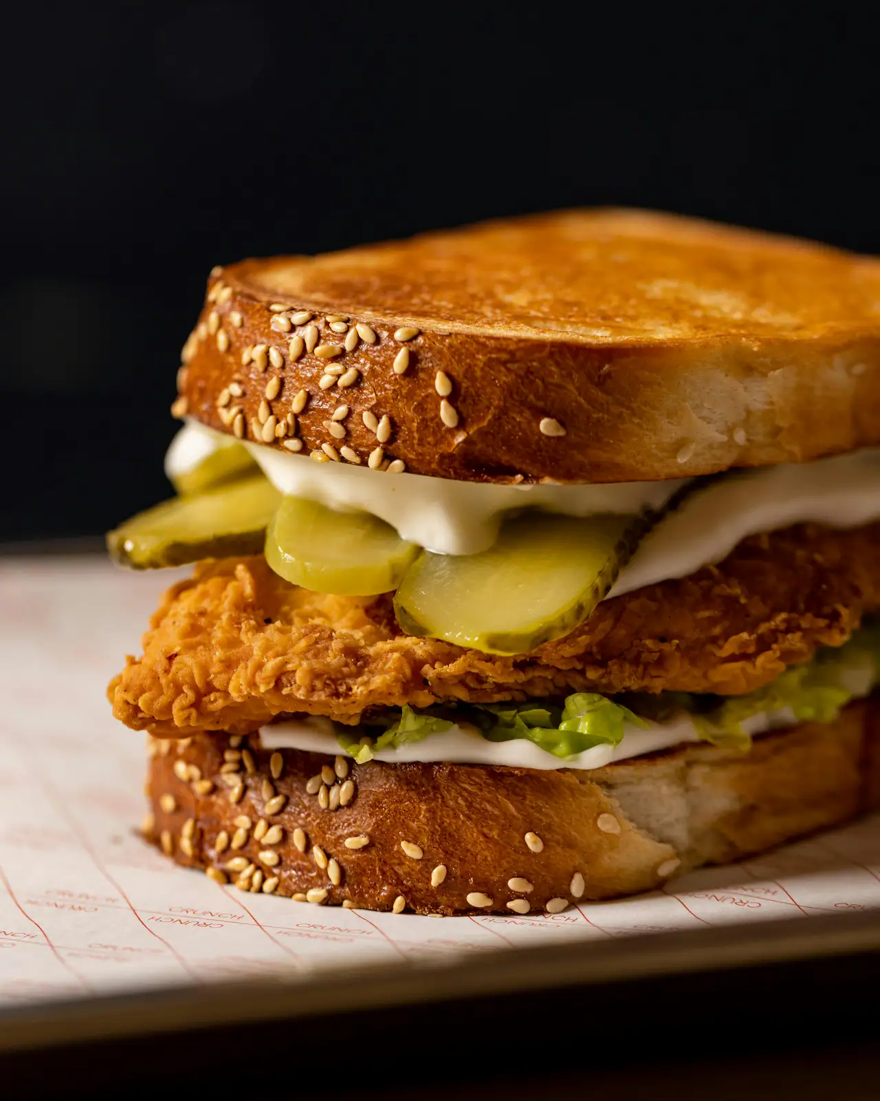 A toasted sesame-topped sandwich with a golden fried chicken cutlet, pickles, mayonnaise and lettuce against a dark background