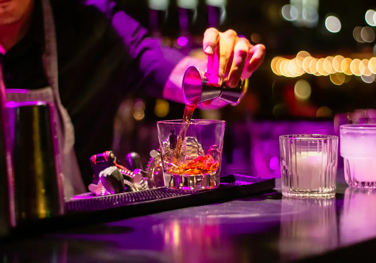 A bartender's hand pouring whisky from a steel jigger into a rocks glass on a dark bar, lit in purple with blurred warm lights behind