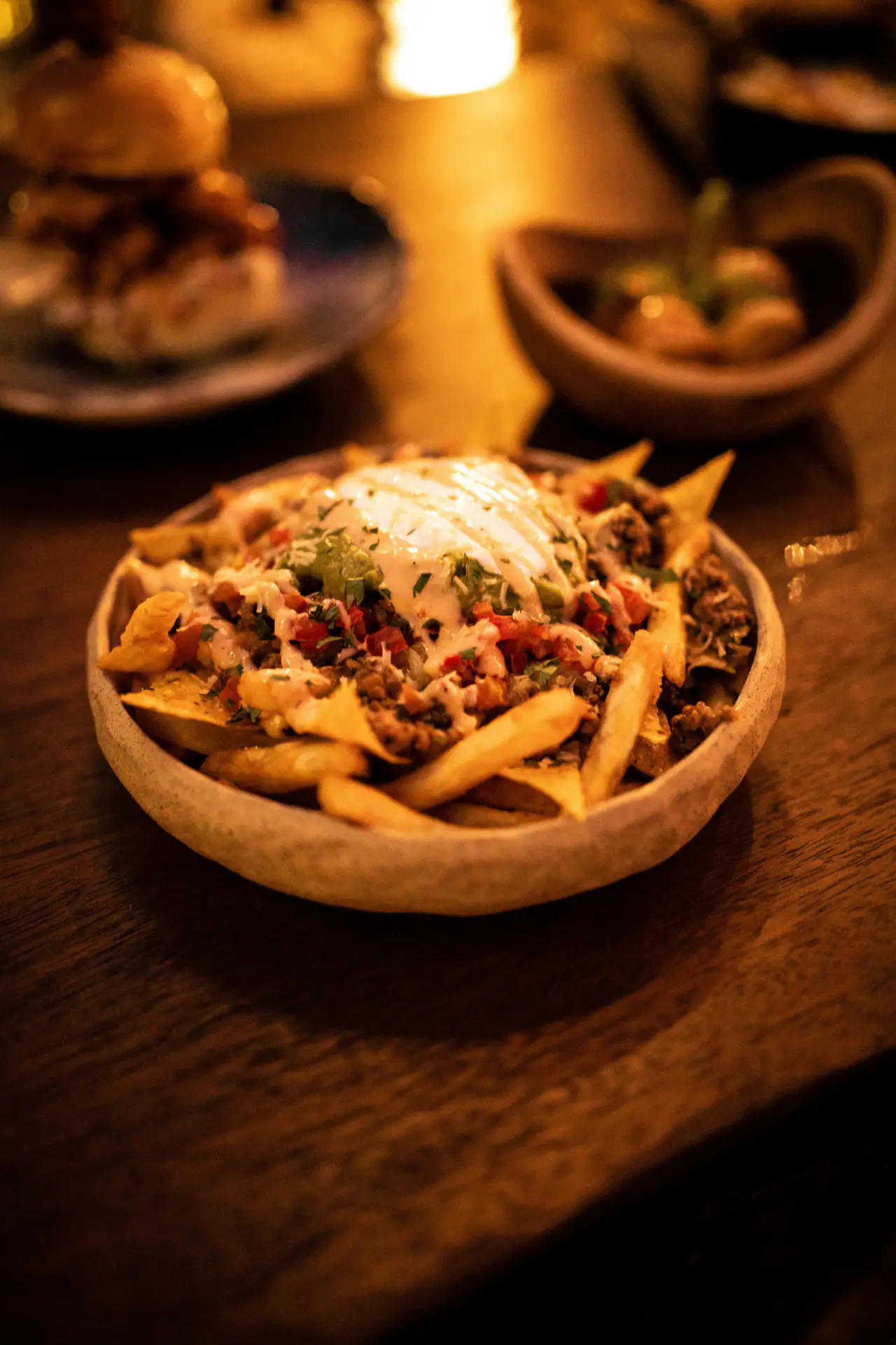 A rustic ceramic bowl of loaded fries topped with sauce, herbs and minced meat on a dark wooden table in warm candlelight