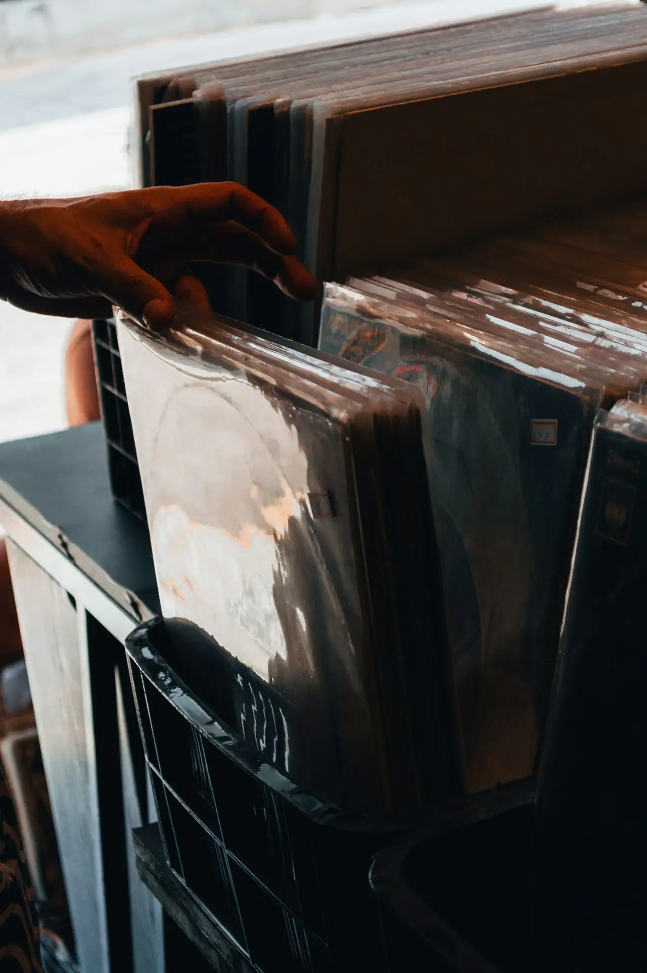 A hand flipping through vinyl LPs in plastic sleeves, standing upright in a black crate in warm daylight