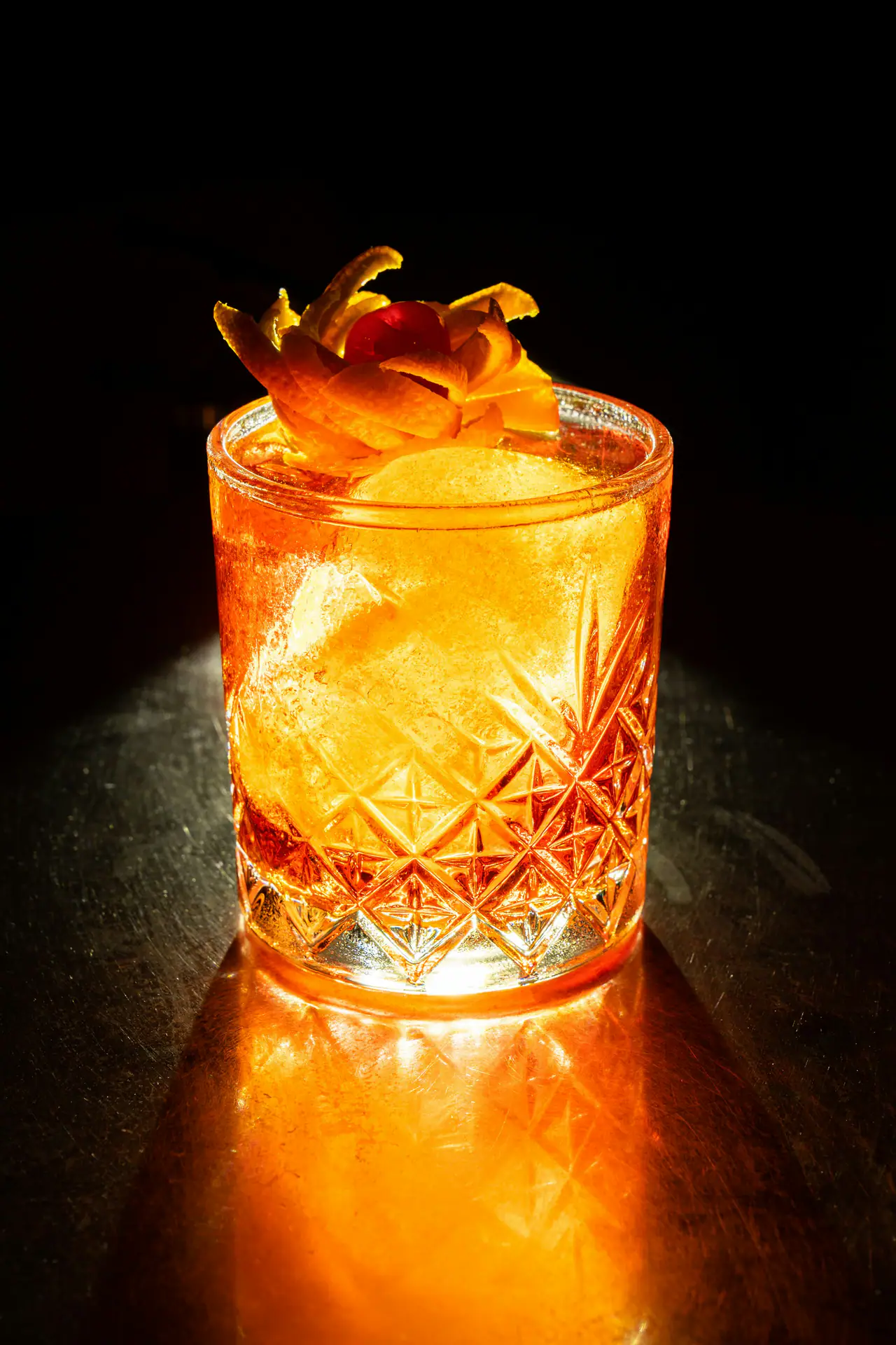 An orange-red cocktail over ice in a cut-crystal tumbler, topped with orange peel and a cherry, lit dramatically against a dark bar top