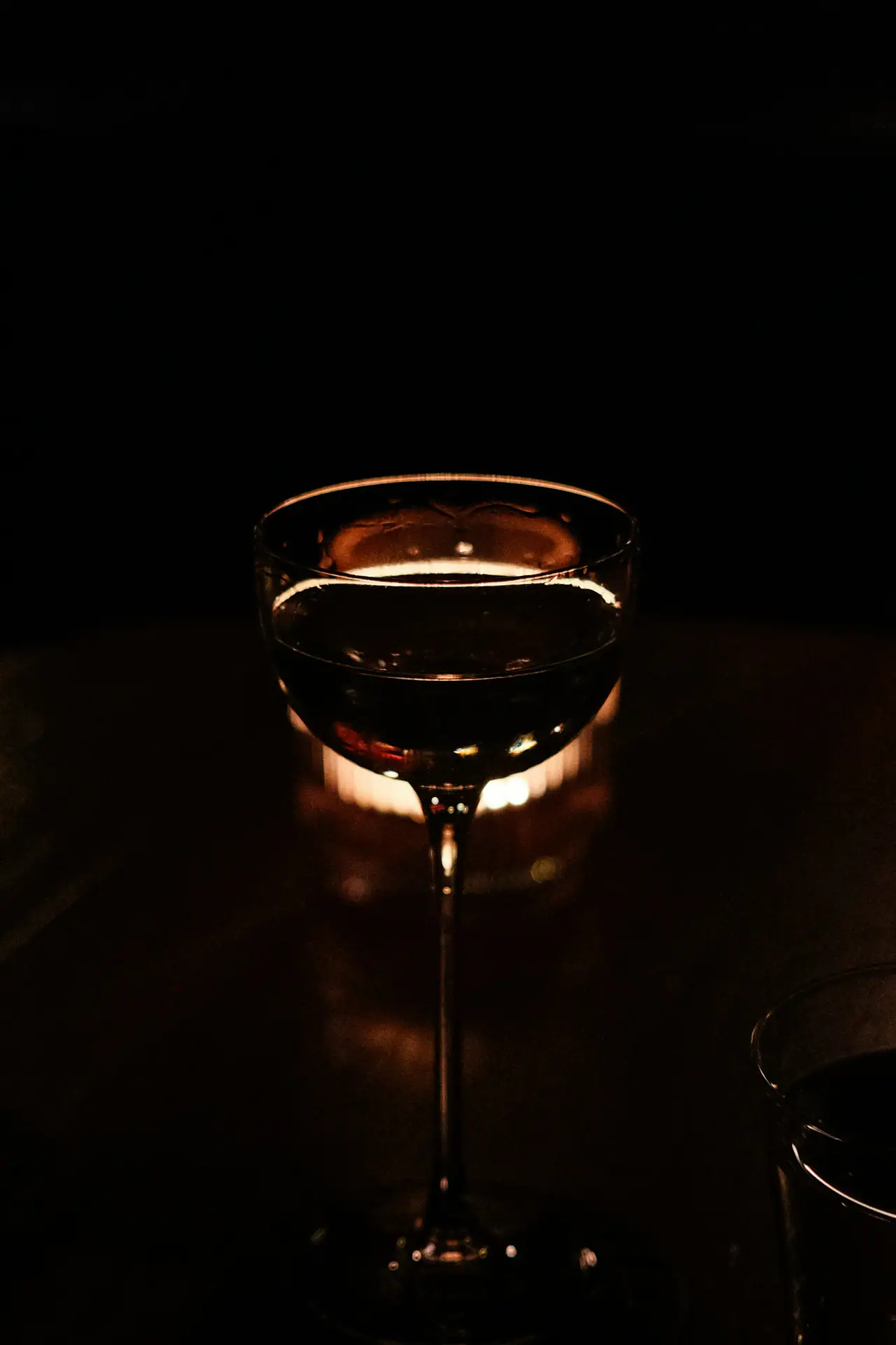 A stemmed cocktail glass backlit by a candle on a dark table in a dim room