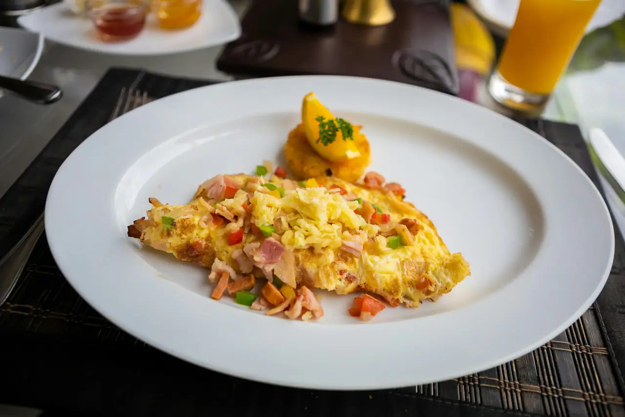 A folded omelette with diced ham, green and red peppers on a white plate, with a glass of orange juice behind