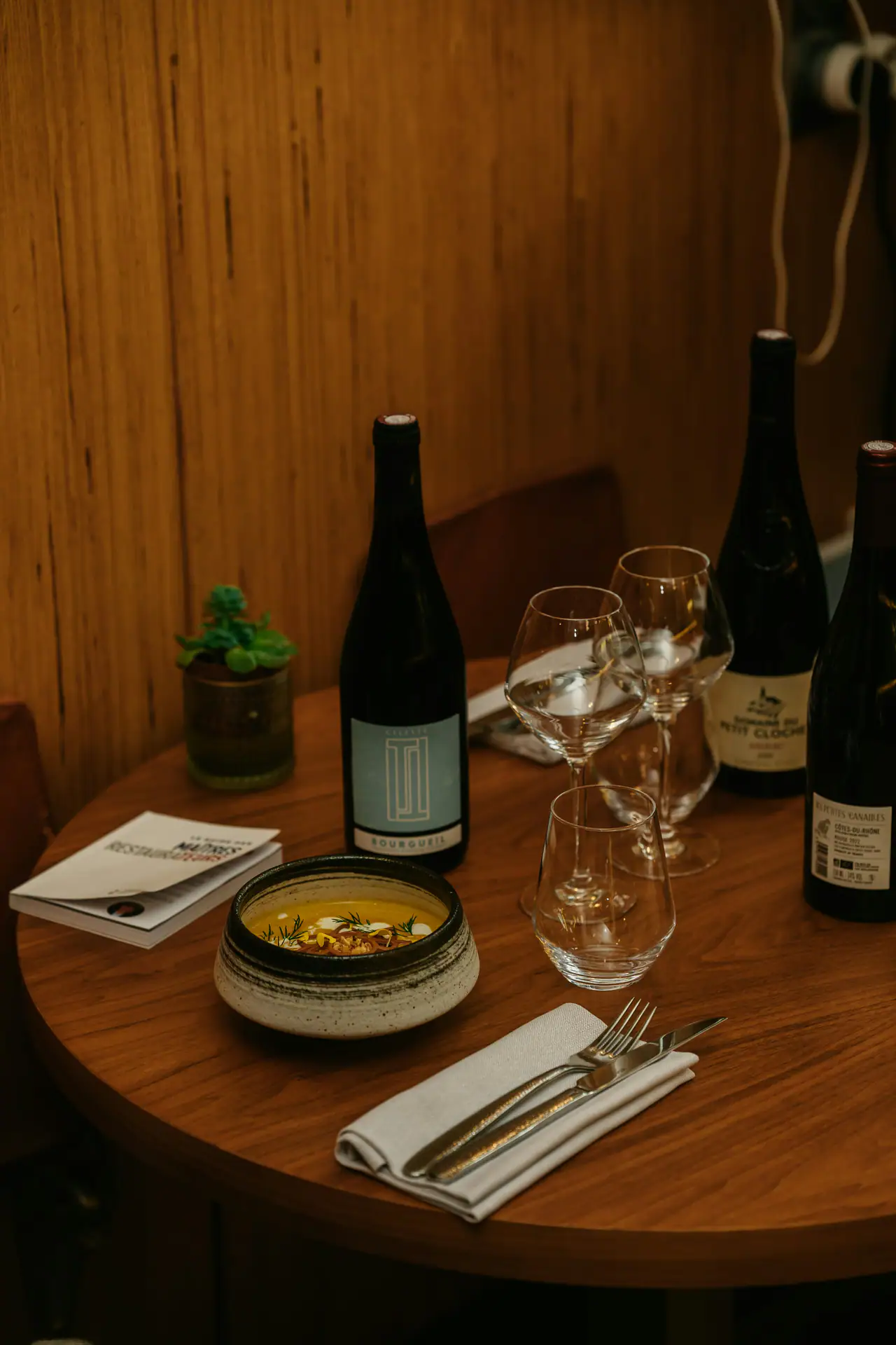 A small round wooden table in a warm, wood-panelled room, set with wine bottles, stemmed glasses, a folded napkin with cutlery and a stoneware bowl of soup