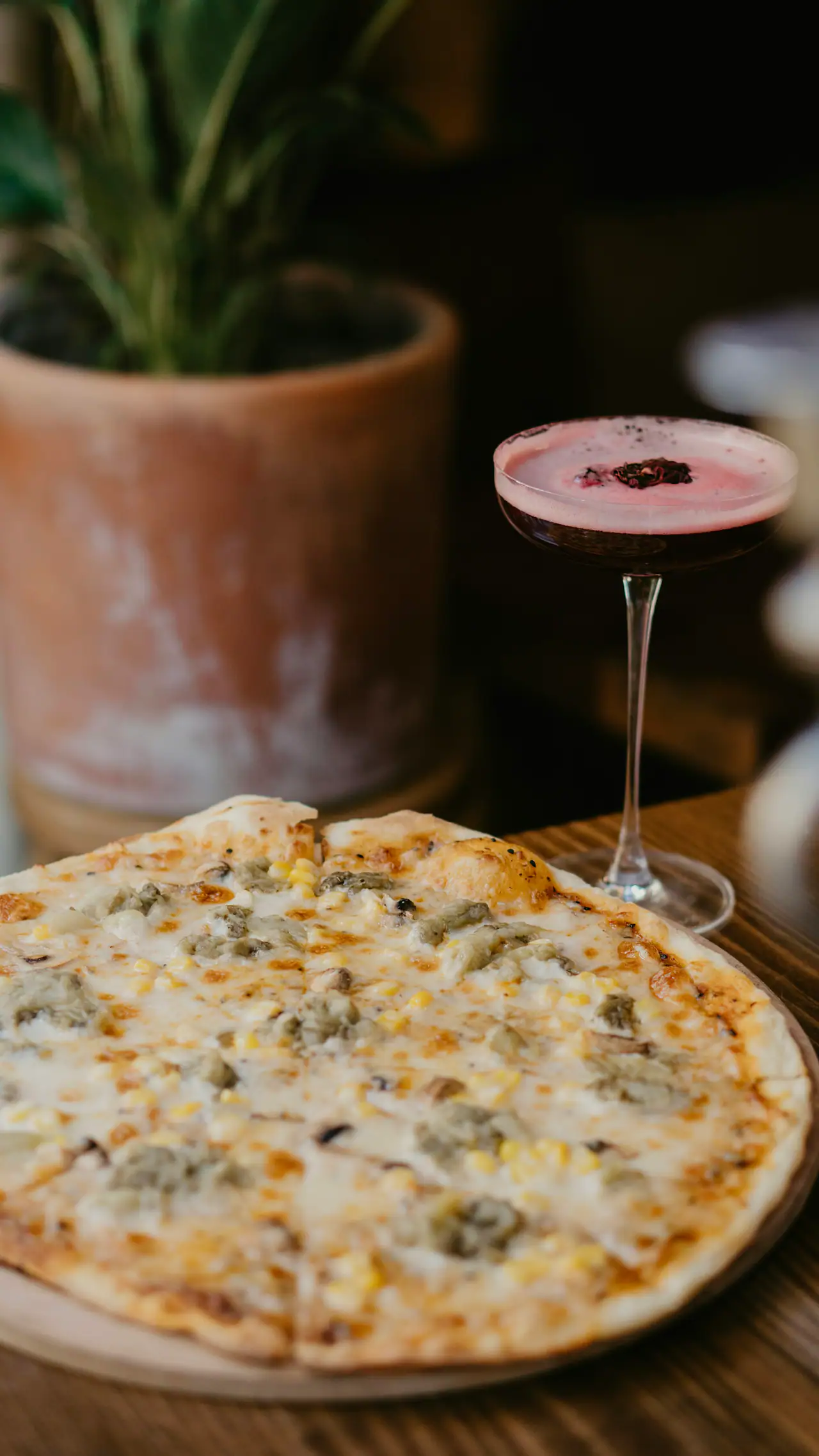 A thin-crust white pizza on a wooden board beside a stemmed coupe glass holding a pink-topped cocktail, with a terracotta planter in the soft dark background