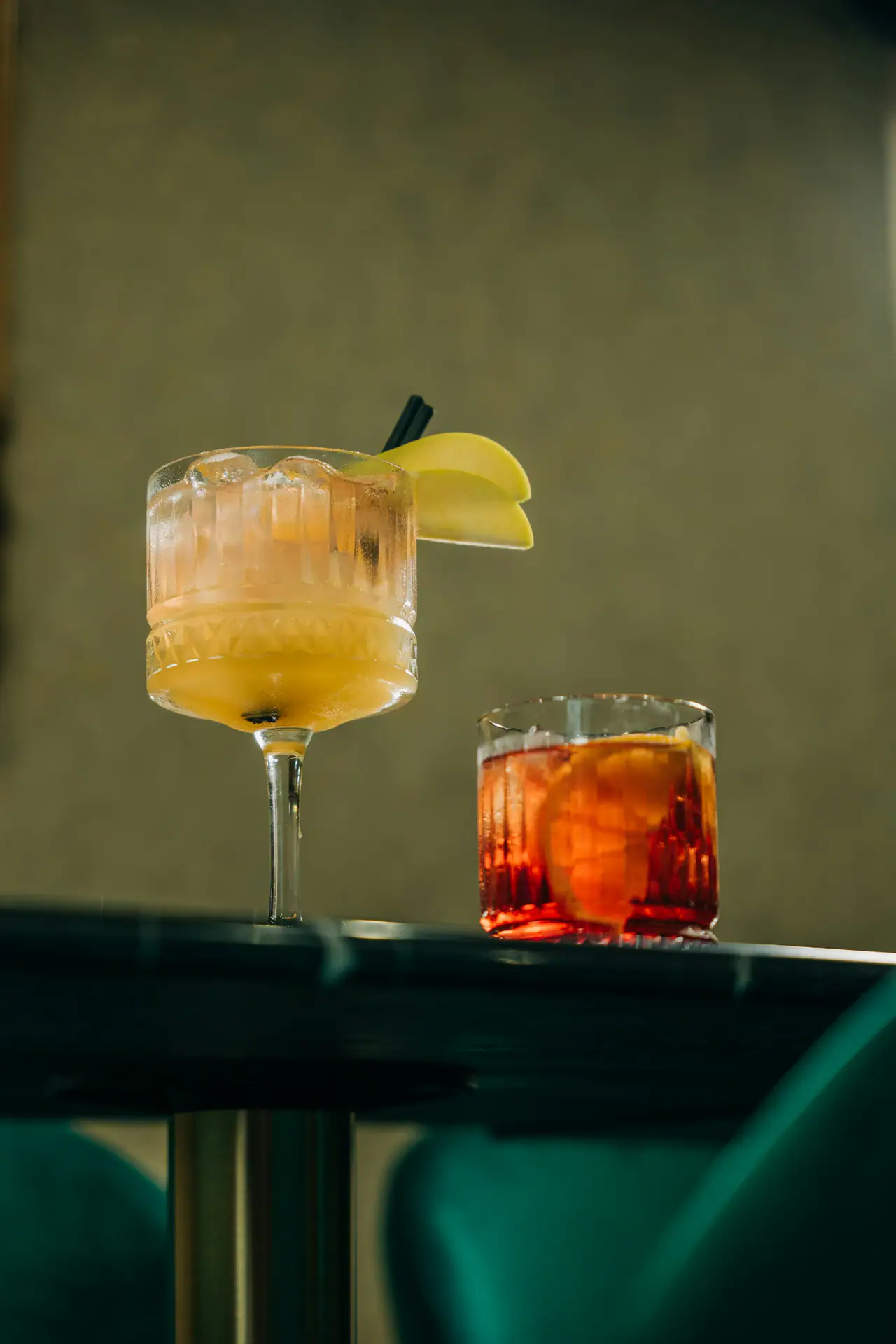 Two cocktails on a dark table in a dim room with green velvet seats: a pale yellow drink in a cut-glass coupe with apple slices, and a red amber drink with citrus in a rocks glass