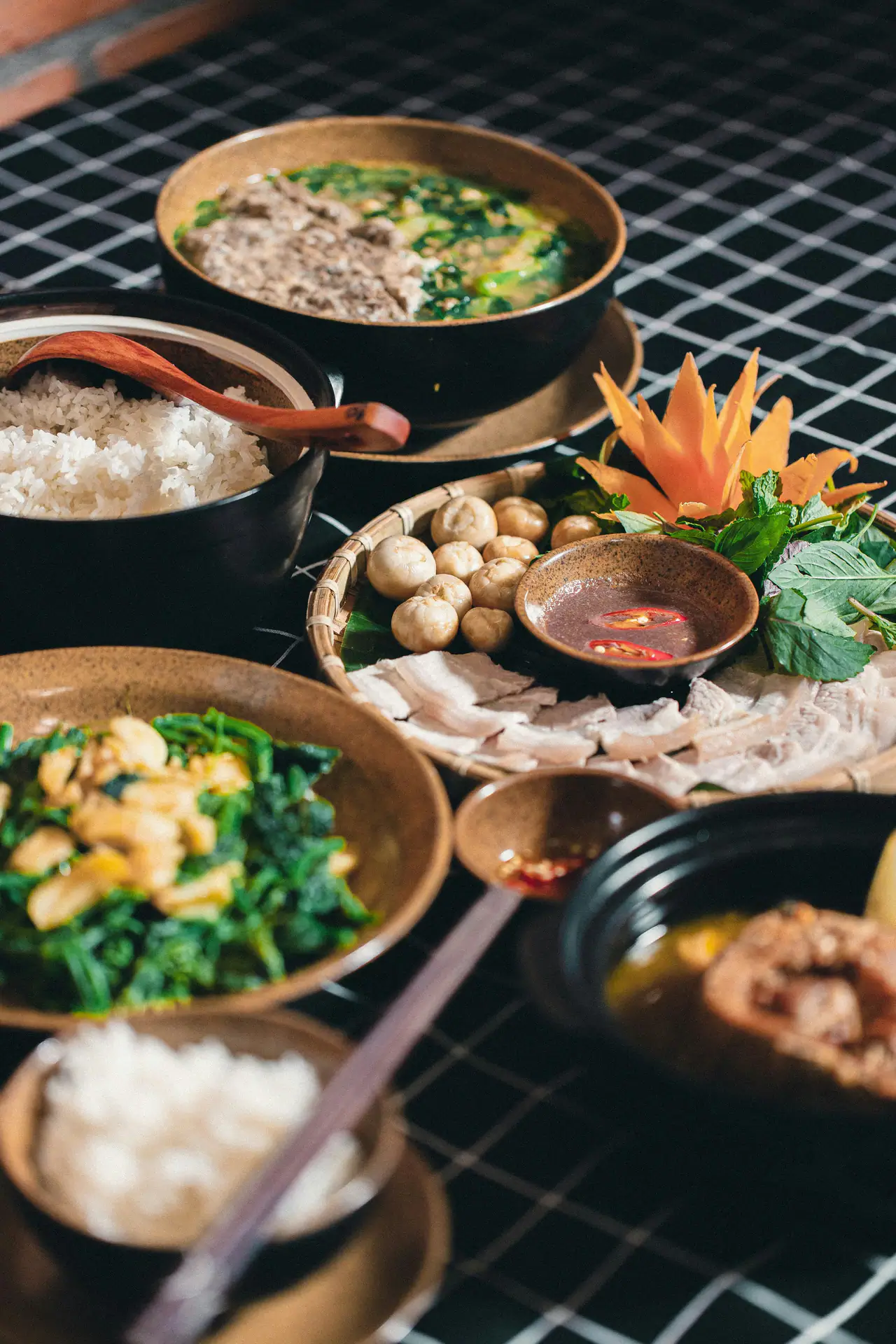 A dark table with shared Vietnamese dishes in black and clay bowls: steamed rice, sliced pork with dipping sauce and herbs, sautéed greens and a green soup