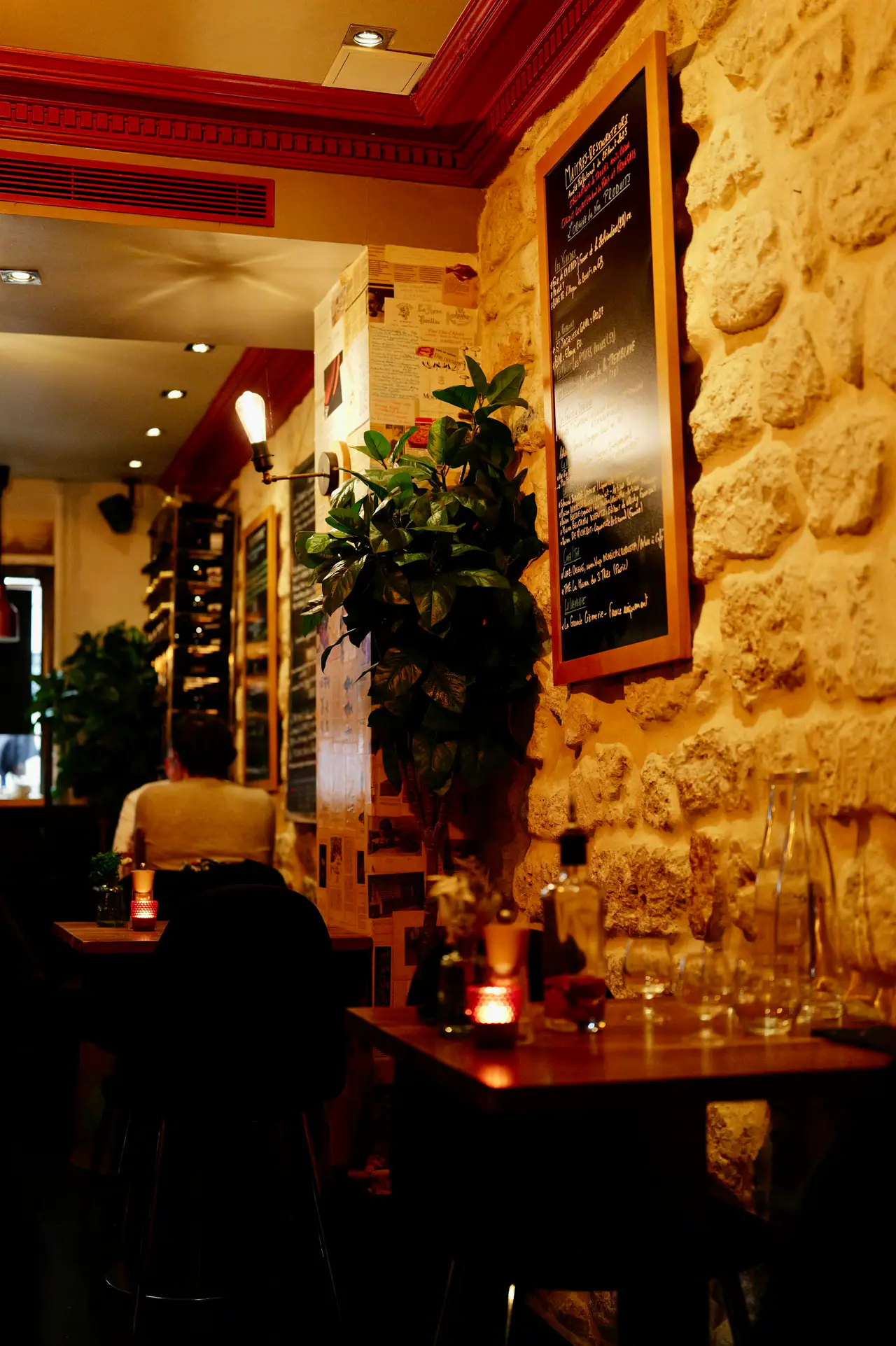 A dimly lit bistro interior with a rough stone wall, a framed chalkboard menu, a candle glowing on a wooden table and wine glasses nearby