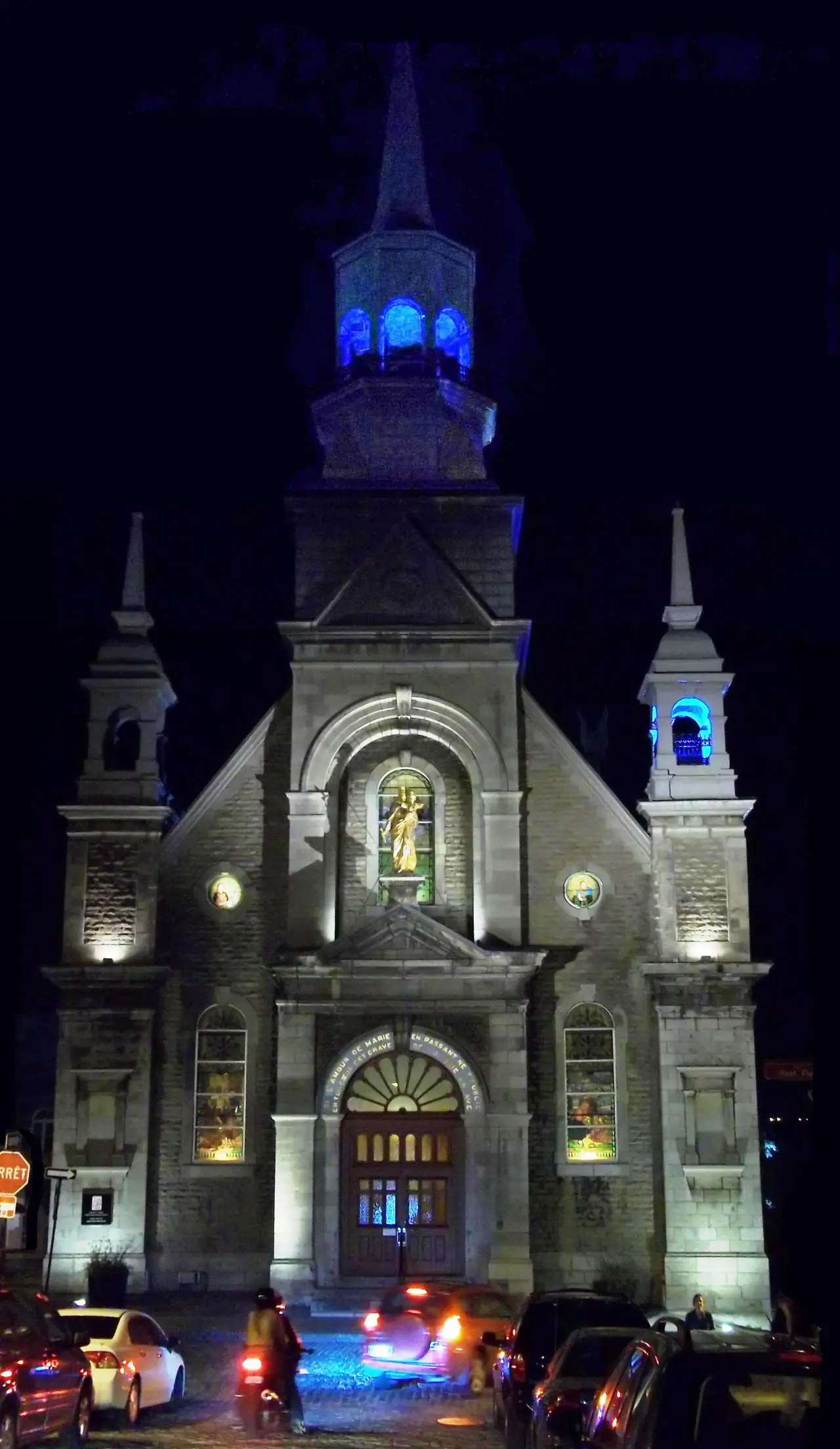 Night view of the stone facade of Notre-Dame-de-Bon-Secours Chapel in Old Montréal, floodlit, with a gilded statue of Mary above the door and blue-lit bell towers against a black sky