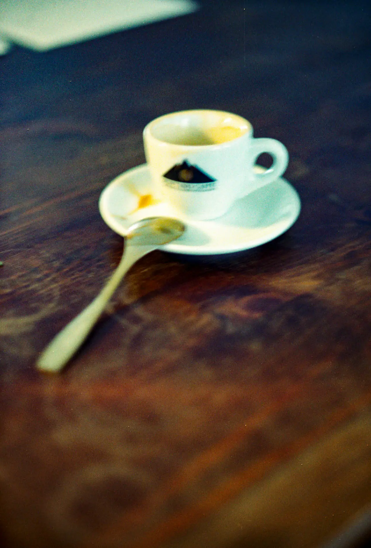 A small white espresso cup and saucer with a spoon resting on a dark, worn wooden table, shot with a soft film-like blur