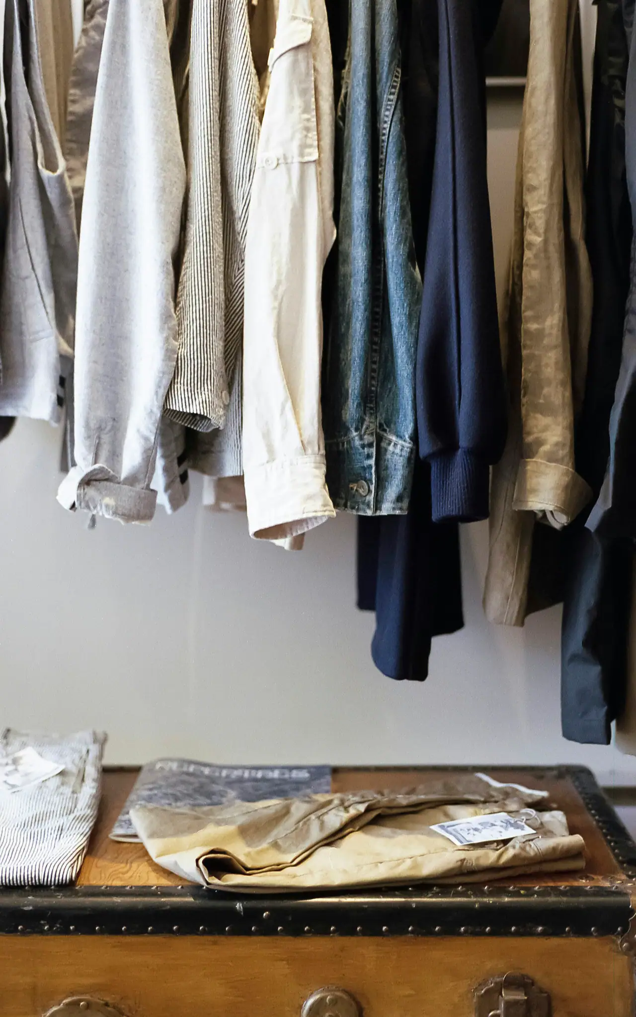 Shirts, a denim jacket and knitwear hanging above a worn wooden trunk holding folded chinos and striped trousers