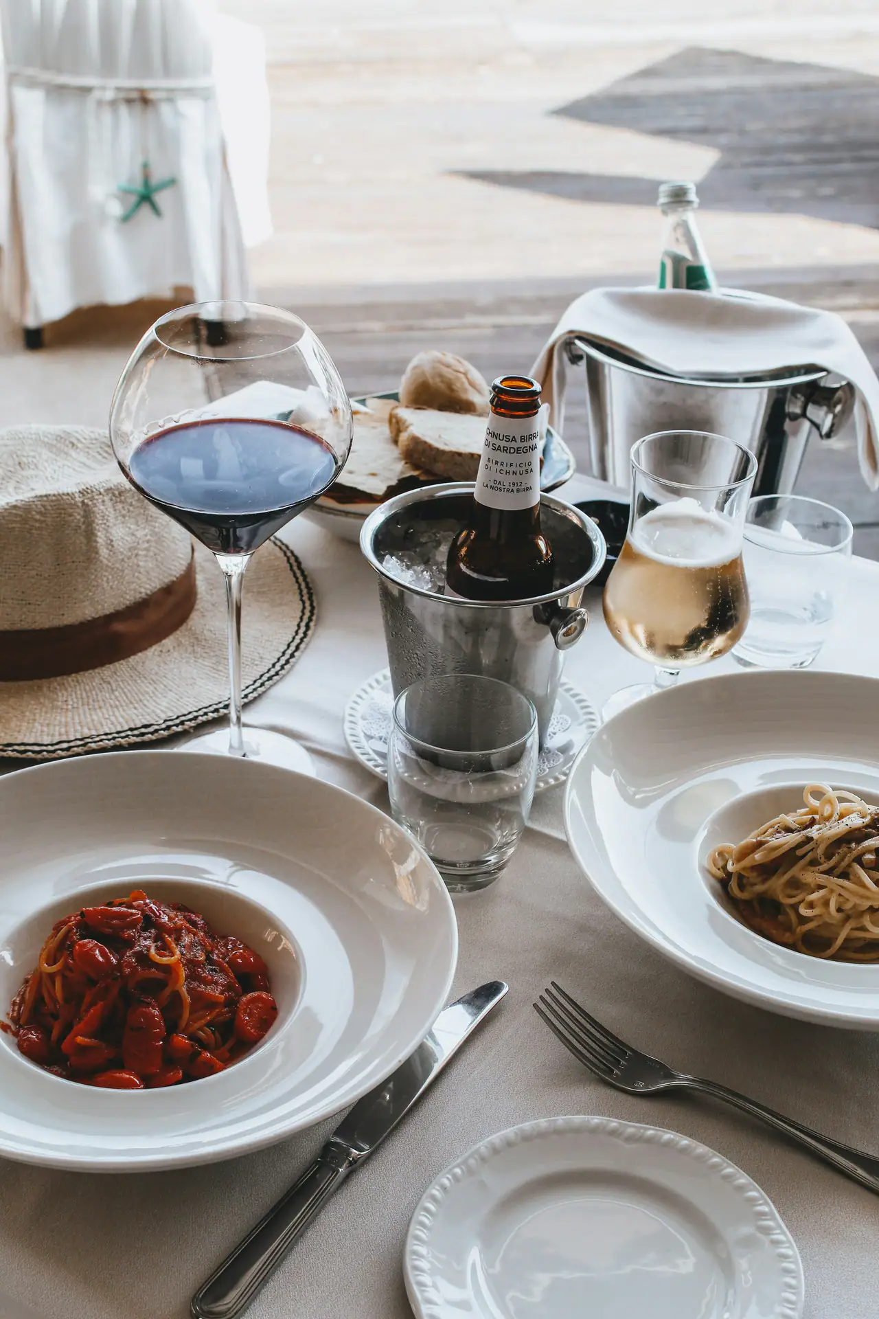 Two bowls of pasta on a white linen-covered table with a glass of red wine, a champagne bucket and a glass of rosé, bread and a straw hat beside them