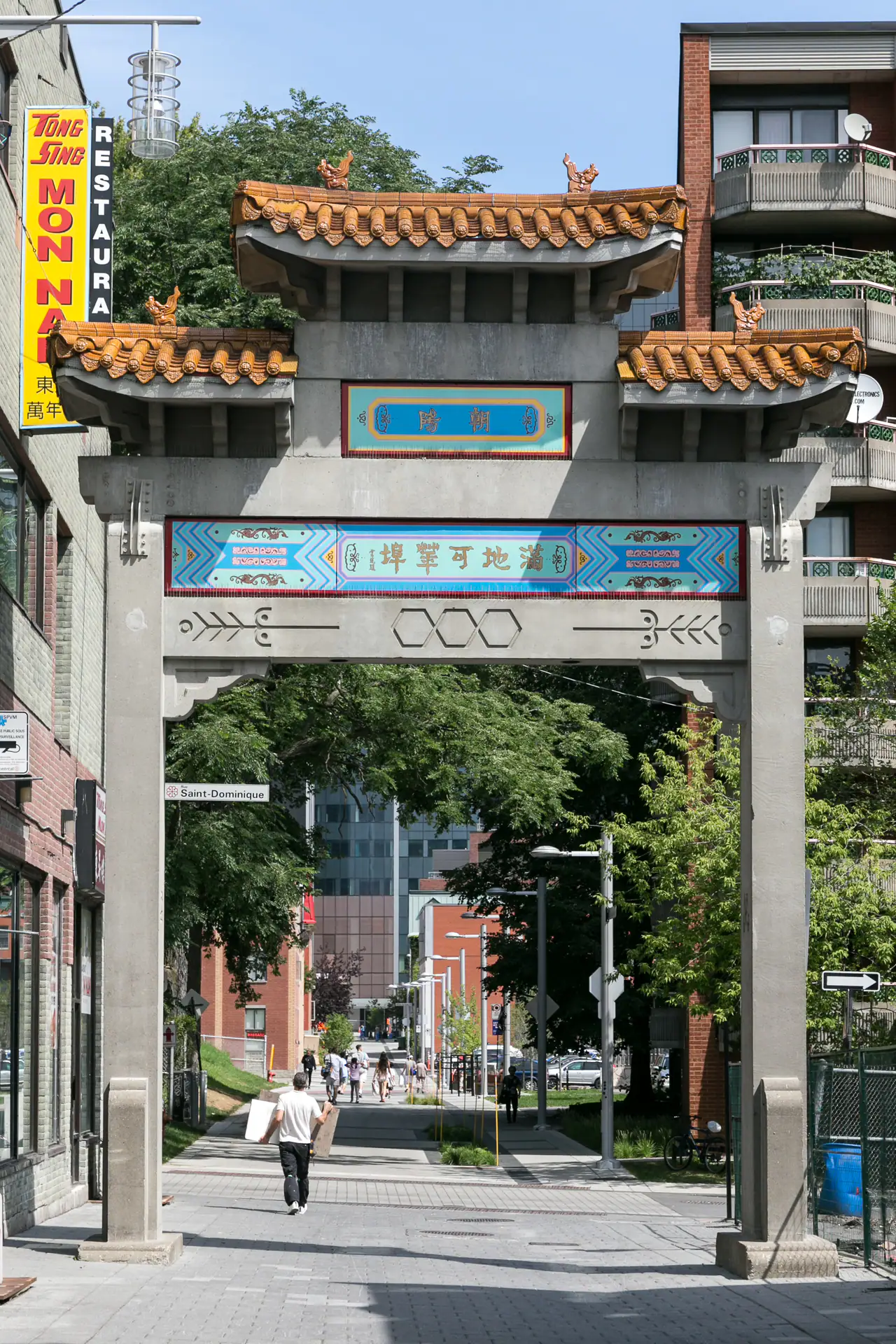 A tiled-roof paifang gate with blue and gold painted panels spans a pedestrian street in Montréal's Chinatown, with trees and brick buildings beyond.