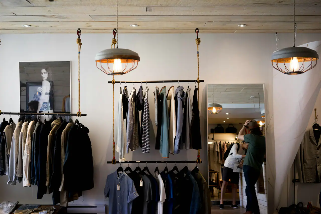 Inside a bright boutique with pendant lamps hung on ropes, shirts and jackets on suspended rails, and a shopper reflected in a tall mirror