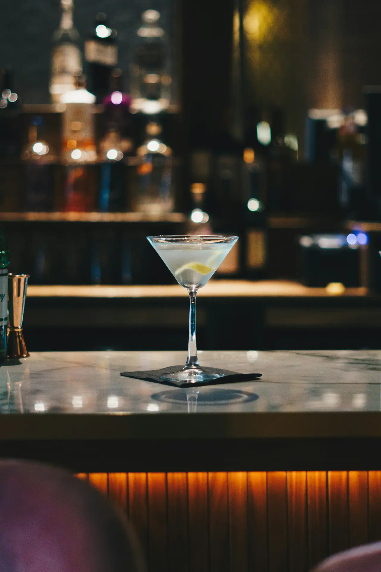 A martini with a lemon twist on a marble bar top, with softly blurred bottles and warm amber lighting behind