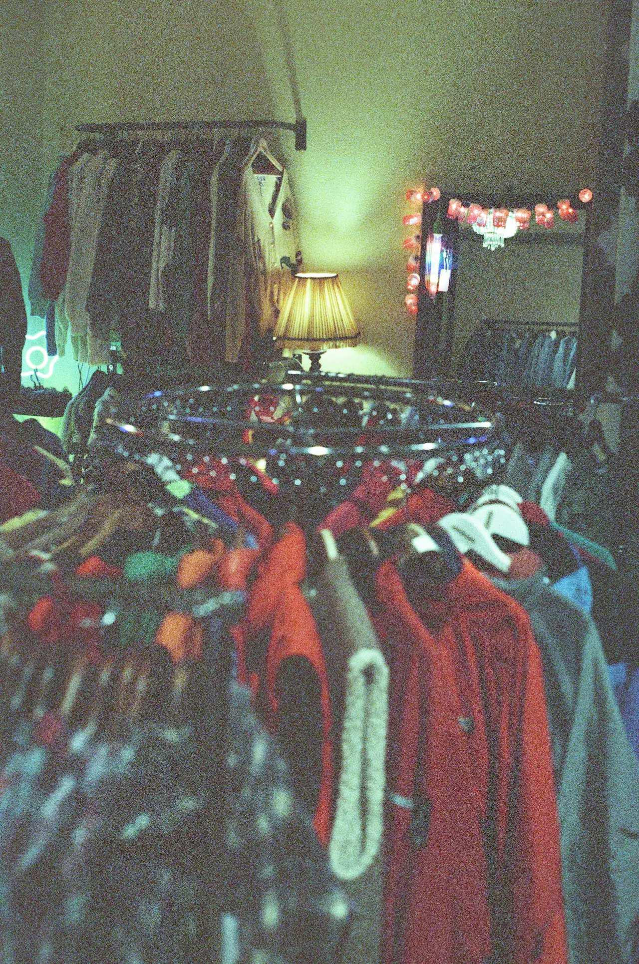 A crowded rail of colourful second-hand clothing in a dim shop, with a glowing lamp, string lights and a mirror in the background