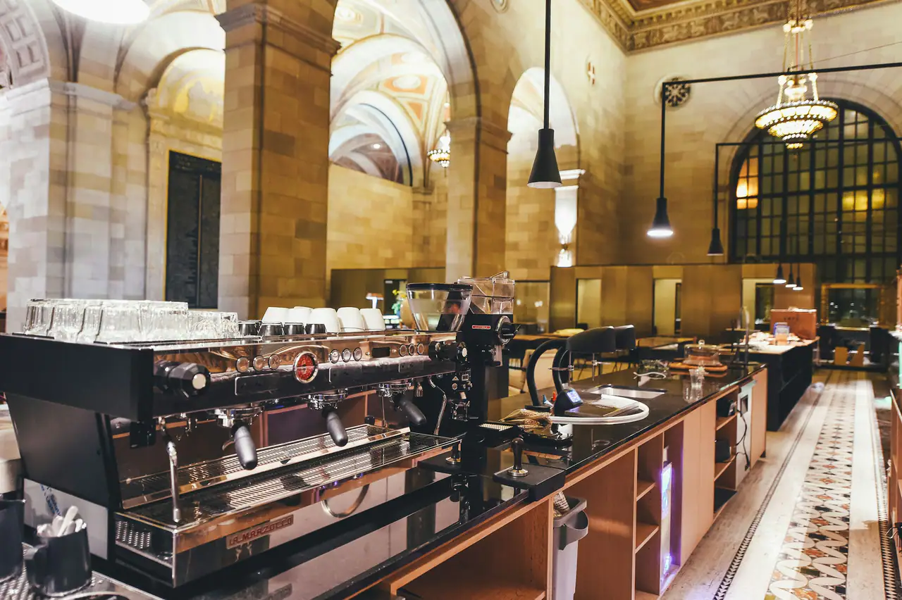 An espresso machine and rows of cups on a counter inside a grand former bank hall with arched, painted ceilings, tall windows and a mosaic marble floor