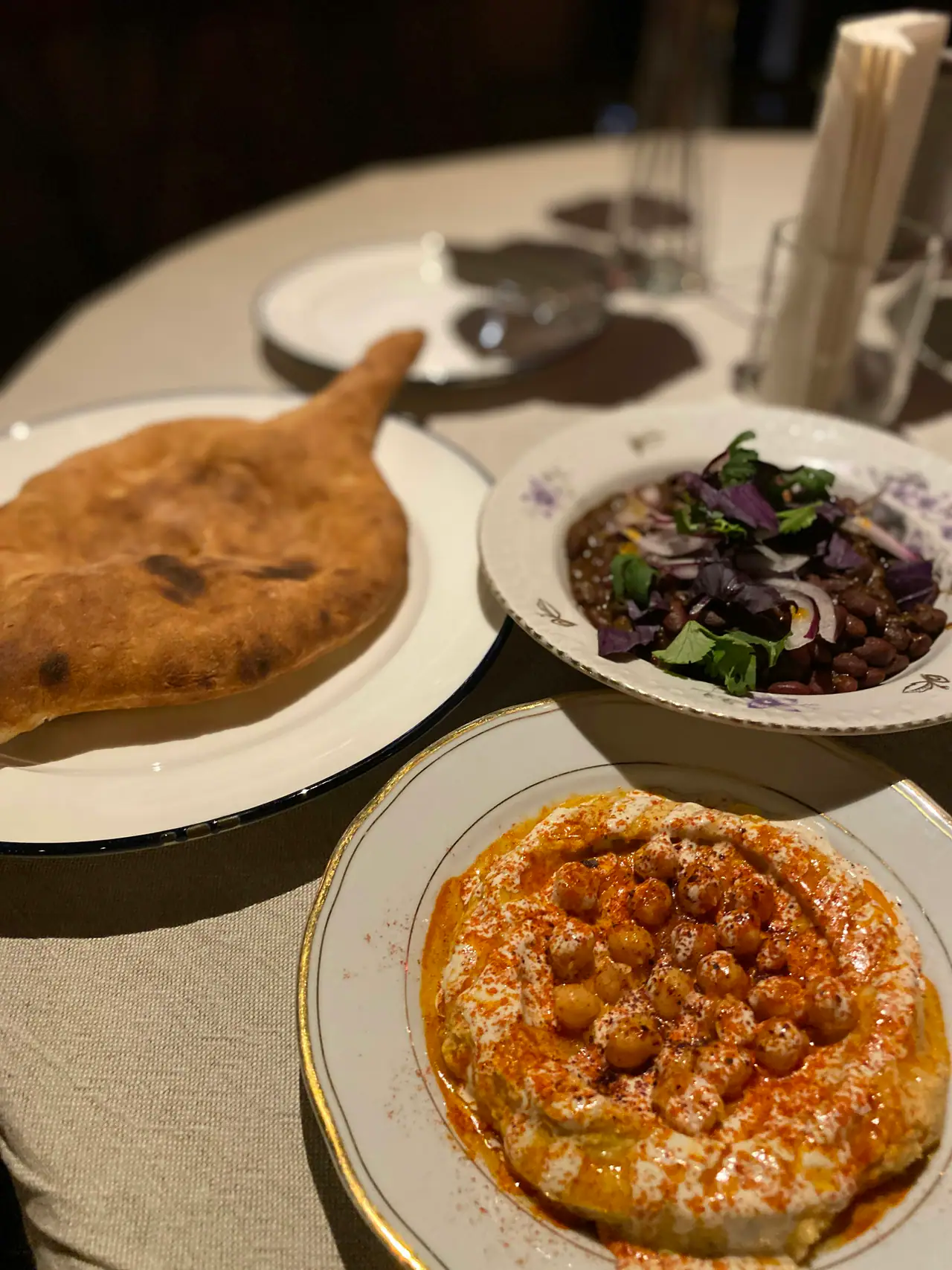 A dim table set with flatbread, a bowl of spiced beans with herbs, and a plate of hummus topped with chickpeas and paprika oil, on linen