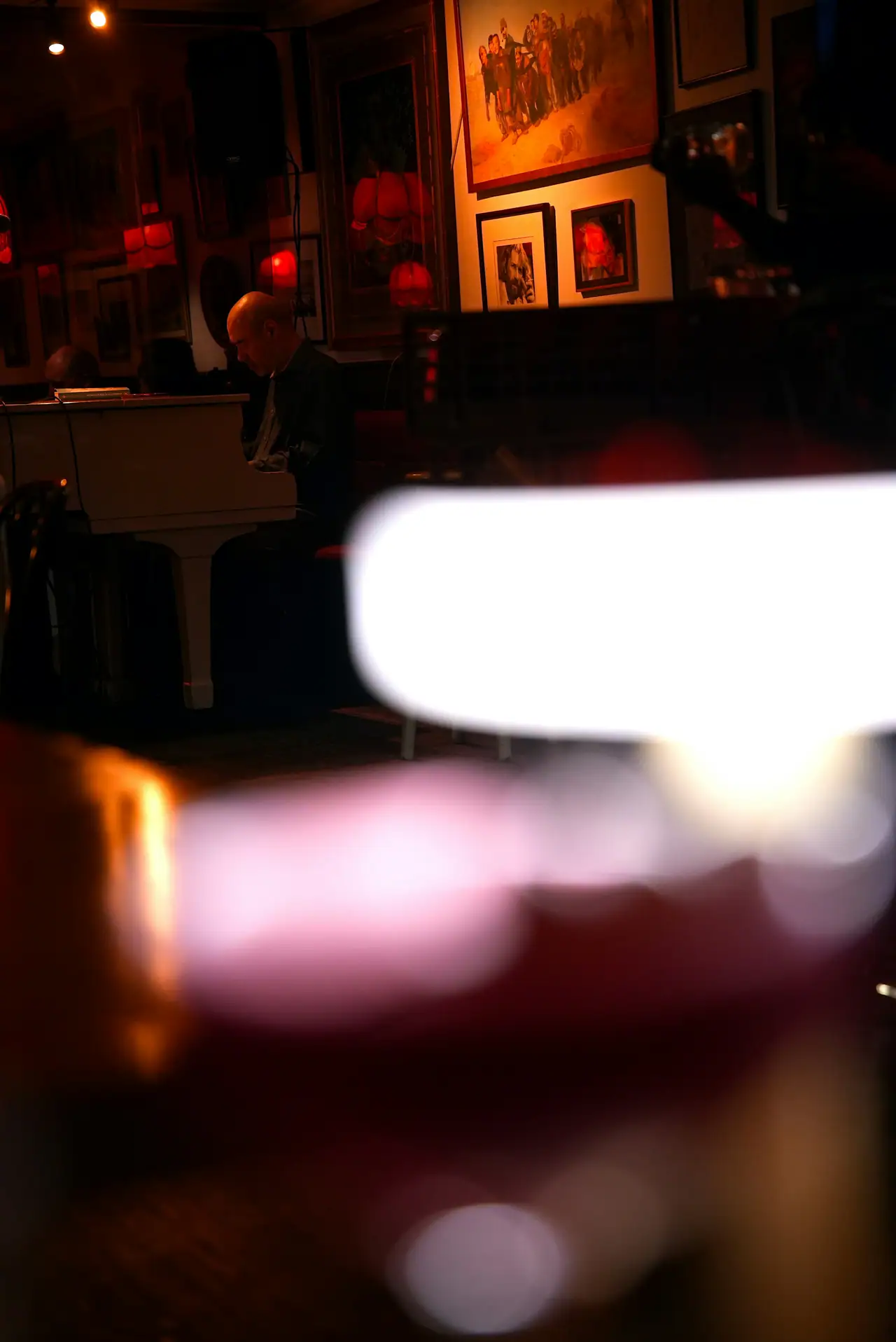 A pianist seated at a white piano in a dim, red-lit room hung with framed paintings, seen past a blurred glass in the foreground. An illustrative jazz-bar scene, not Dièse Onze itself.