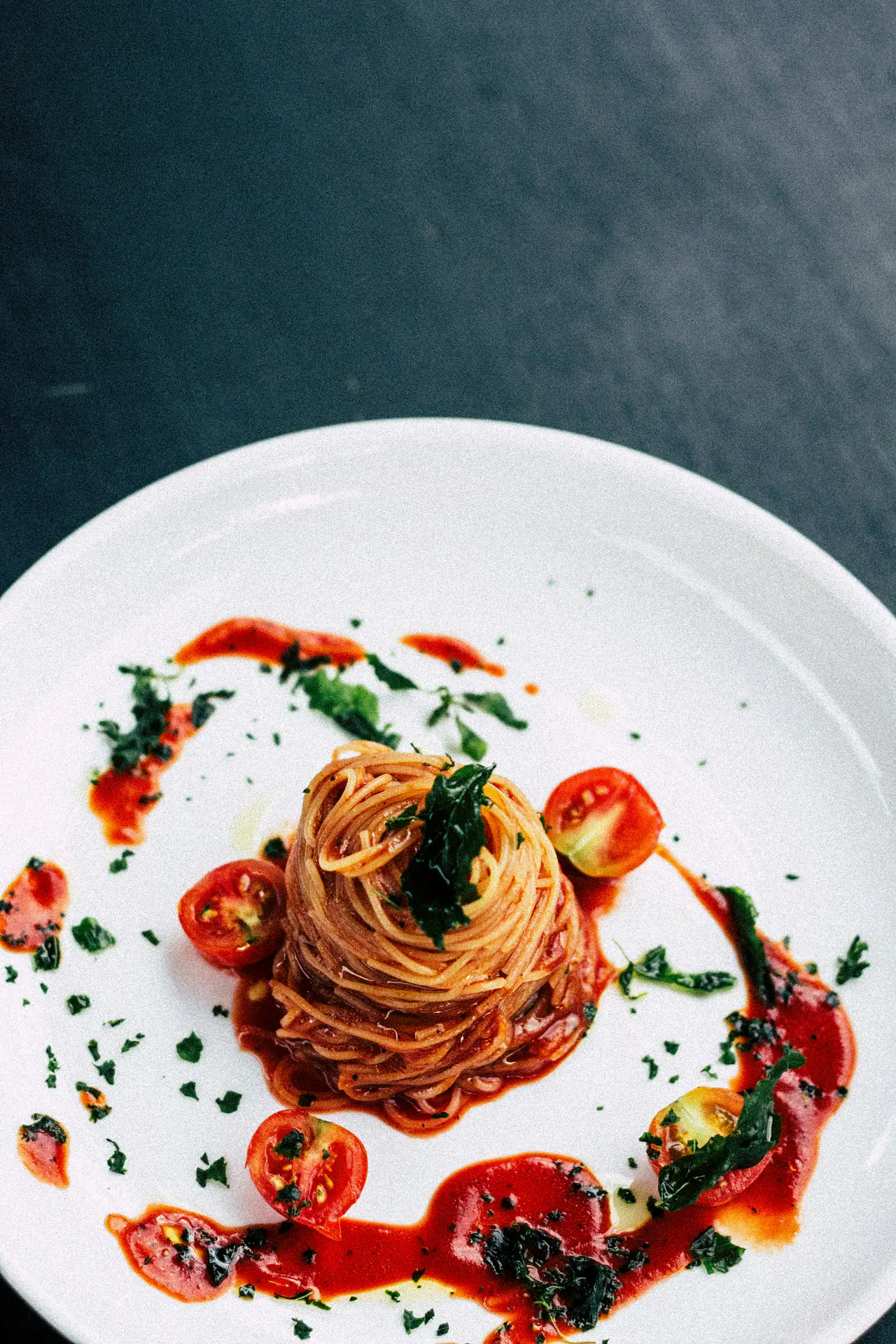 A nest of thin spaghetti in tomato sauce with halved cherry tomatoes and fresh herbs on a white plate against a dark slate surface