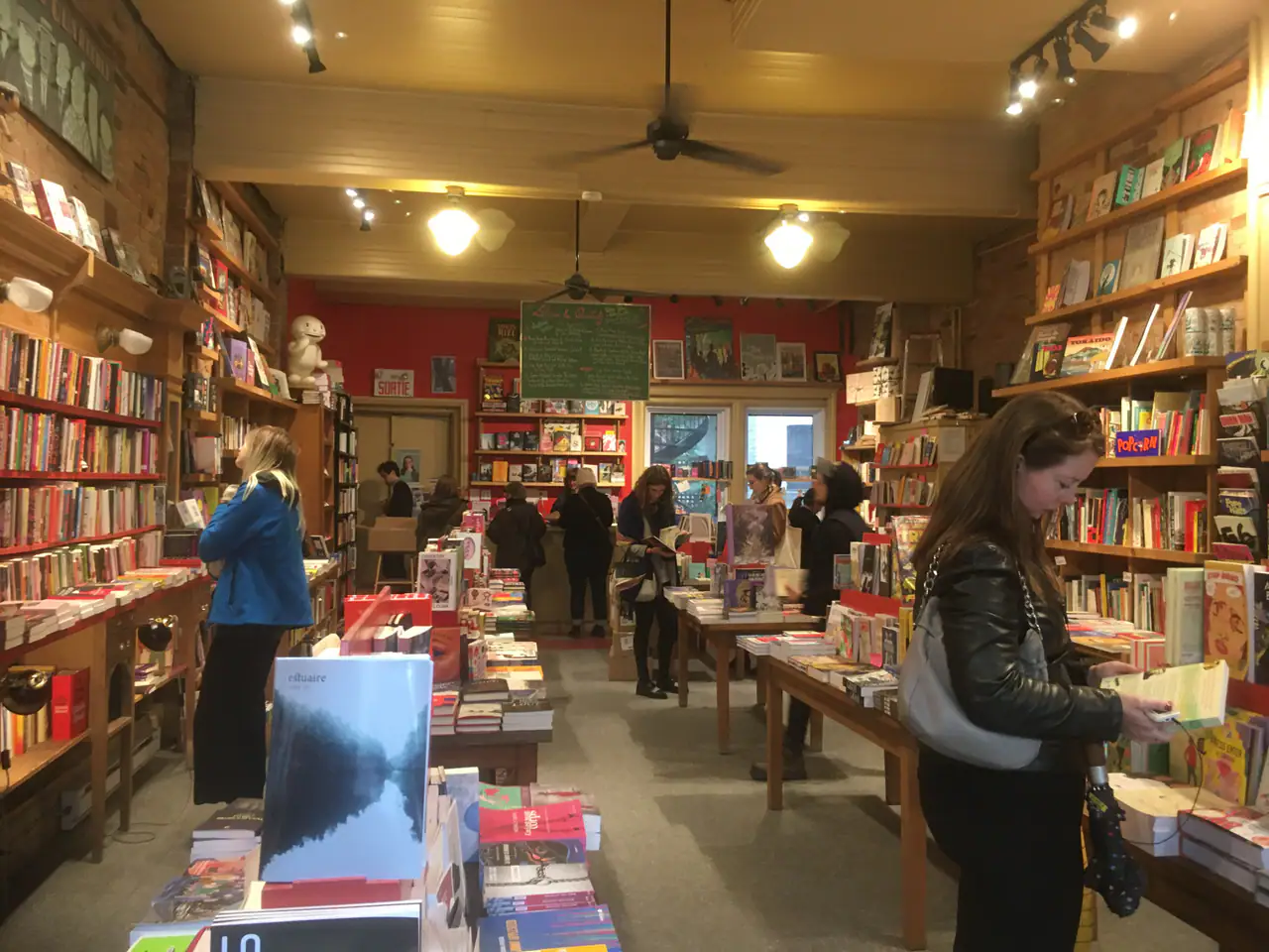 Inside Librairie Drawn & Quarterly in Mile End: customers browsing tables of books beneath exposed brick, wooden shelves and a red back wall