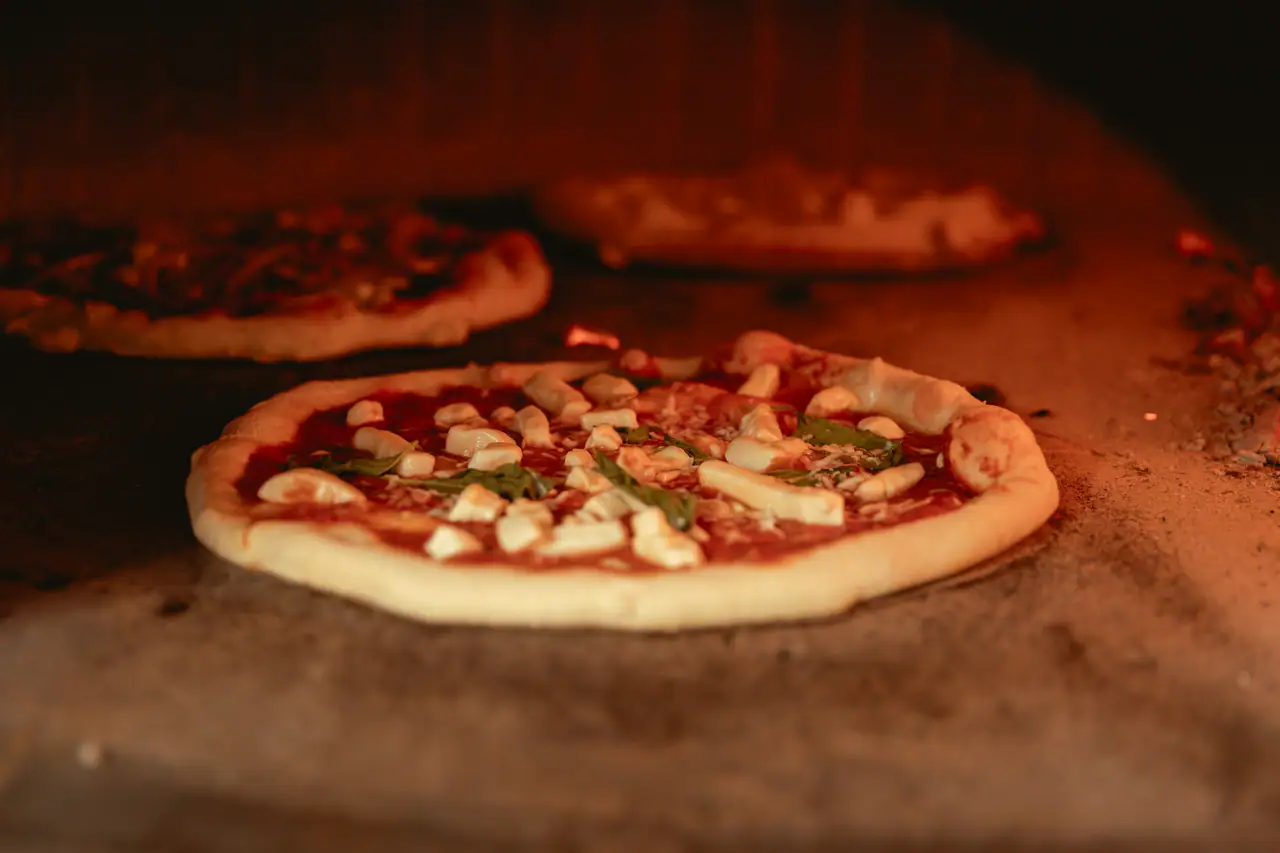 A pizza with tomato, mozzarella and basil baking on the stone floor of a glowing wood-fired oven, with other pizzas blurred behind it