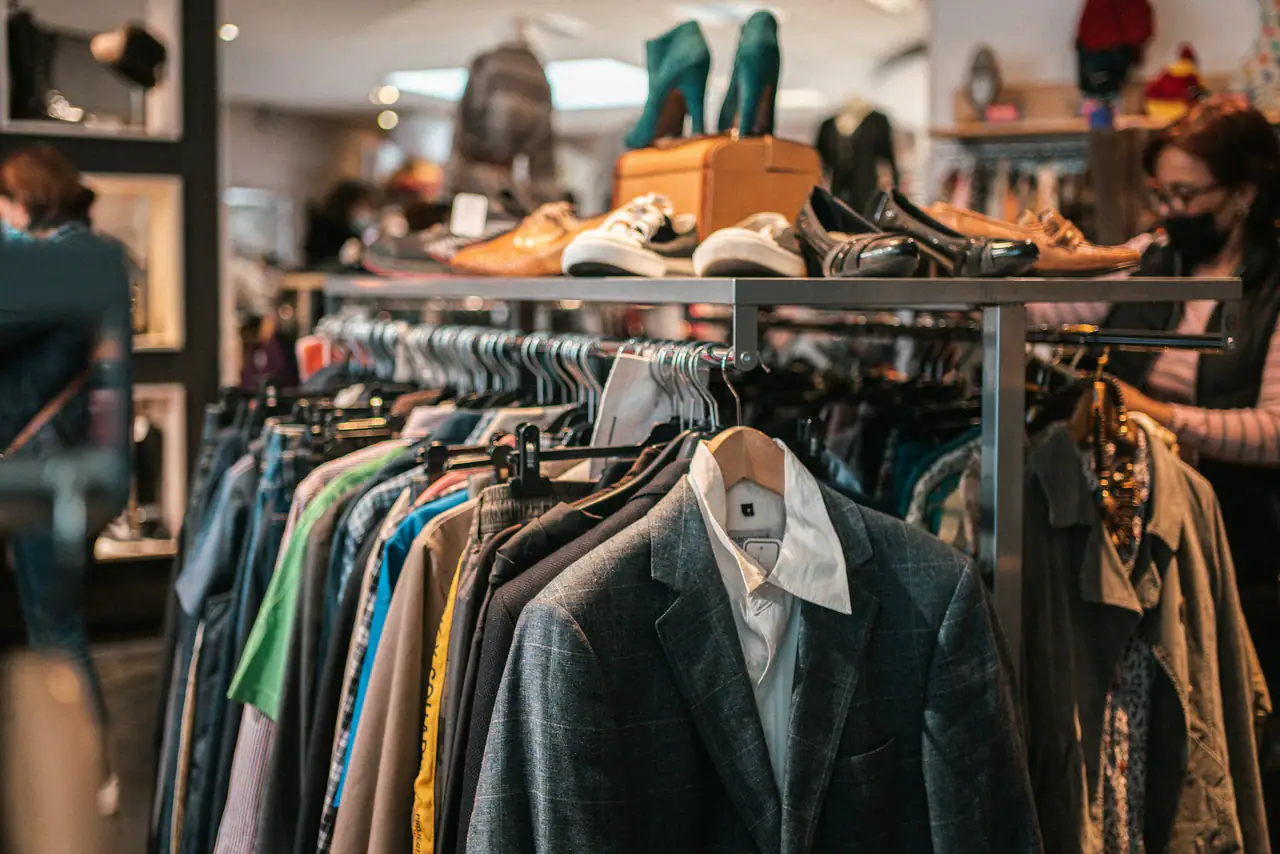 A crowded rail of secondhand jackets and shirts in a bright thrift shop, a grey checked blazer in front and shoes on a shelf behind