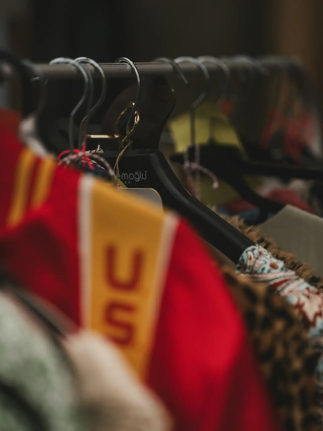 Close-up of a crowded rail of vintage clothes on black hangers, with a red and gold varsity-style knit, leopard print and a patterned fabric in warm, soft focus