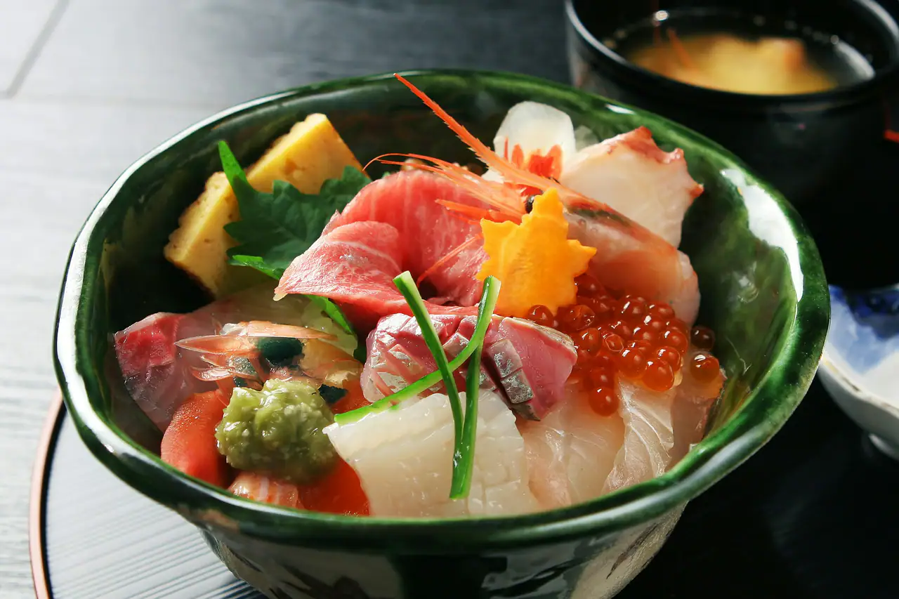 A green glazed bowl of chirashi, assorted sashimi with salmon roe and fresh wasabi over rice, beside a bowl of miso soup on a dark table