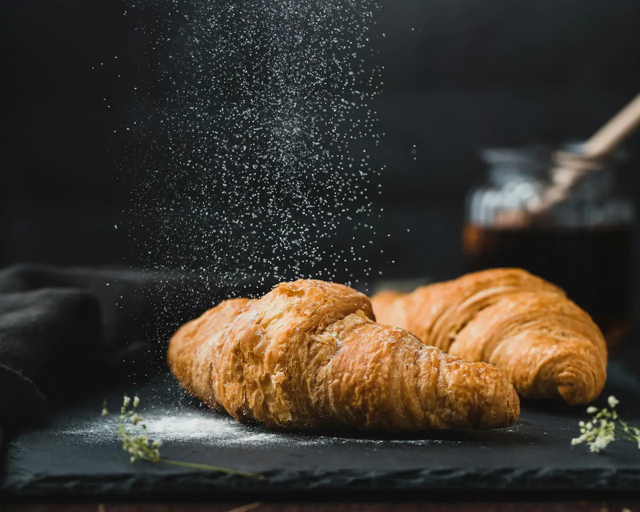 Two golden croissants on a dark slate board with powdered sugar falling over them, a jar of honey behind