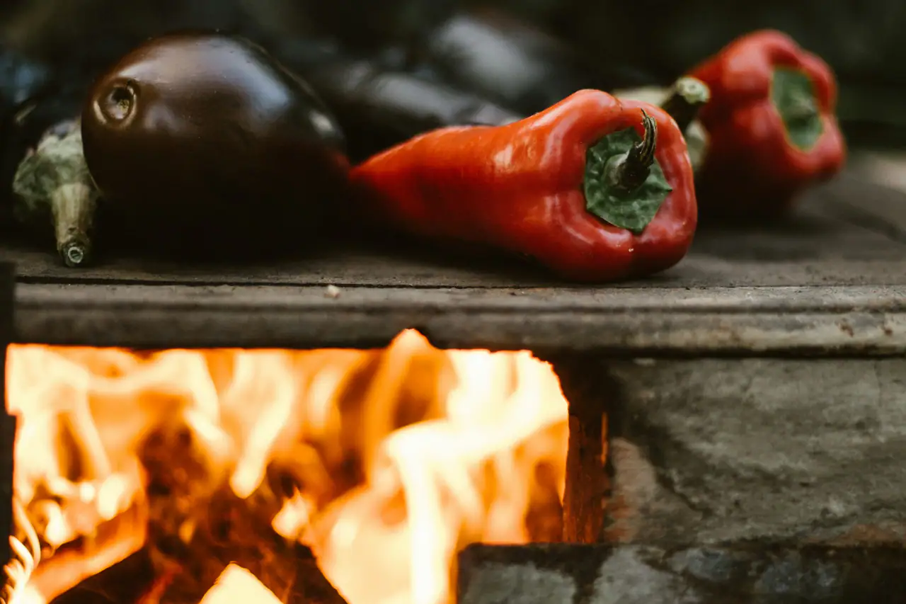 Red peppers and a dark eggplant roasting on a stone ledge above an open wood fire