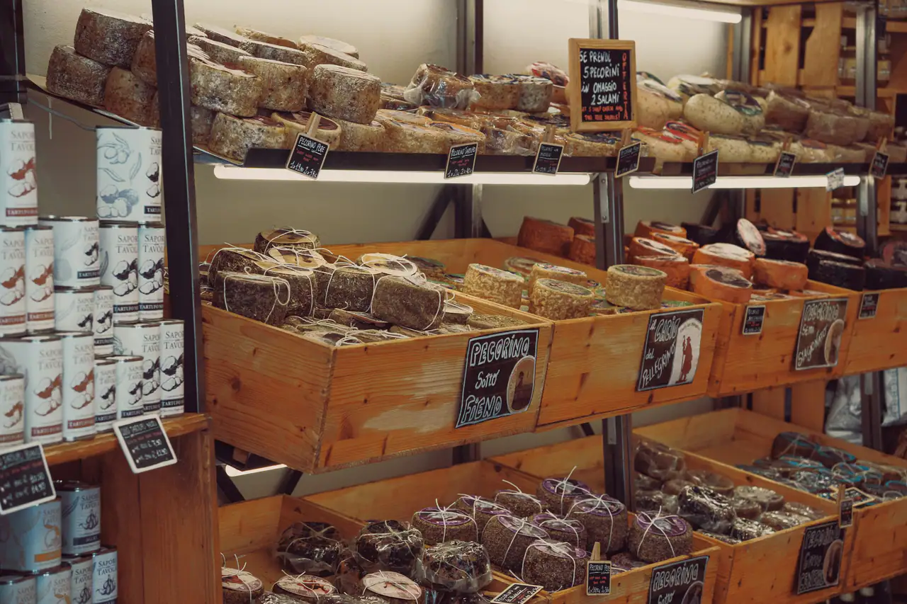 Wooden crates of aged cheese wheels and wedges on shop shelves with small chalkboard labels, beside stacked tins