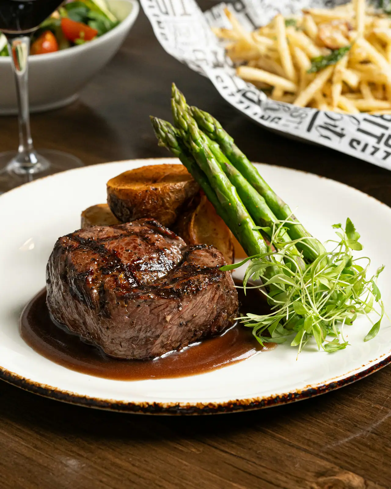 A grilled filet steak in red wine sauce on a white plate with roasted potatoes and asparagus, a glass of red wine behind, on a dark wooden table