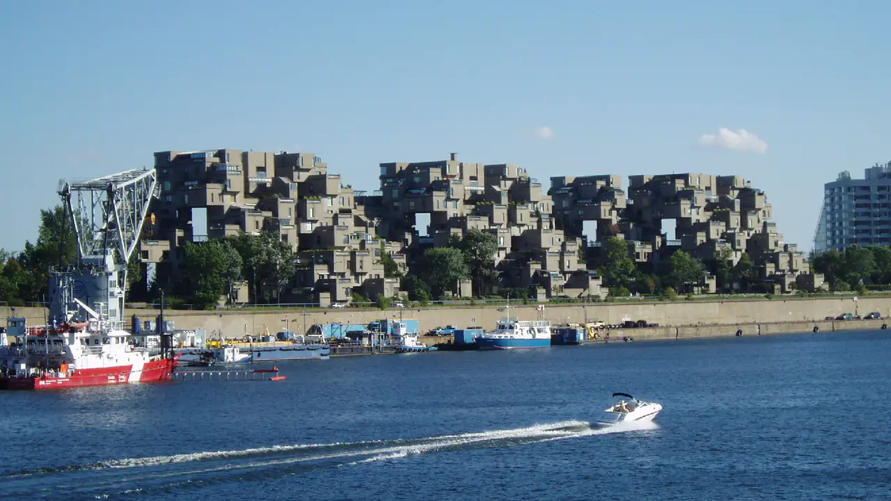 Habitat 67's stacked concrete housing modules seen across the harbour water, with trees on the terraces and a tugboat and small boats in front, under a clear blue sky.
