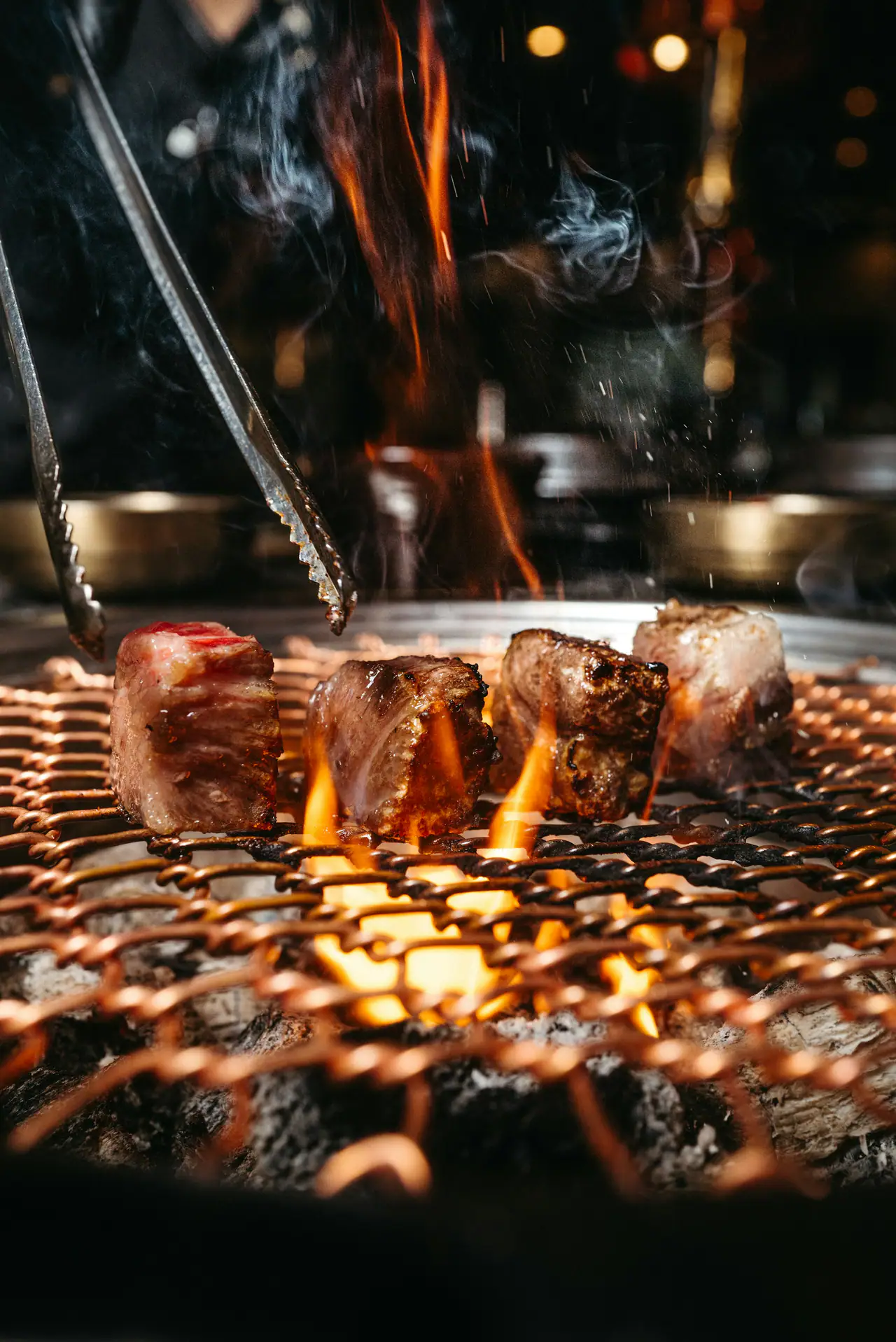 Cubes of beef searing over open flame on a copper-toned mesh grill, with tongs above and a dark restaurant glowing behind