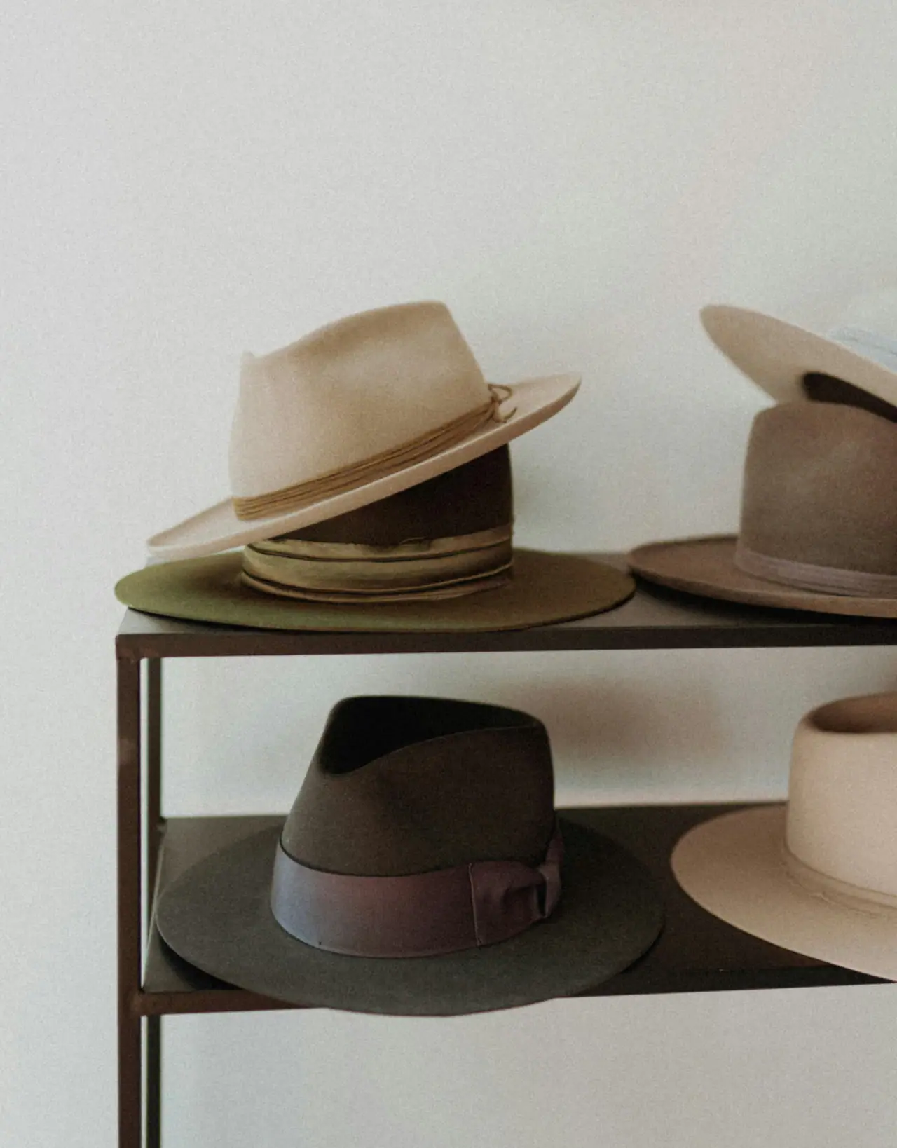 Felt fedoras in cream, olive and brown stacked on a two-tier metal shelf against a plain white wall
