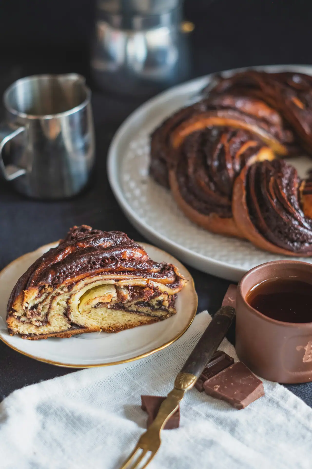 A slice of swirled chocolate babka on a gold-rimmed plate beside the rest of the twisted loaf, with chocolate squares and a linen napkin on a dark table