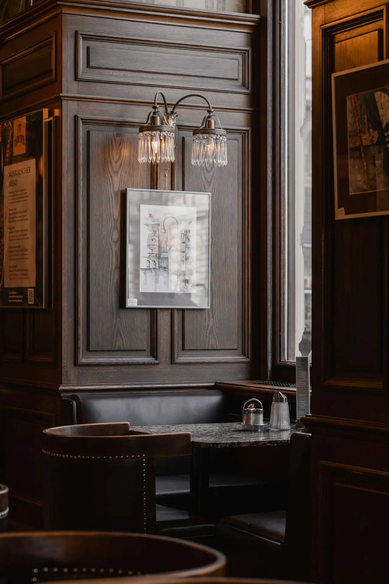 A corner of a dark wood-panelled café-brasserie with a leather banquette, a small marble table and a framed print lit by a crystal wall sconce