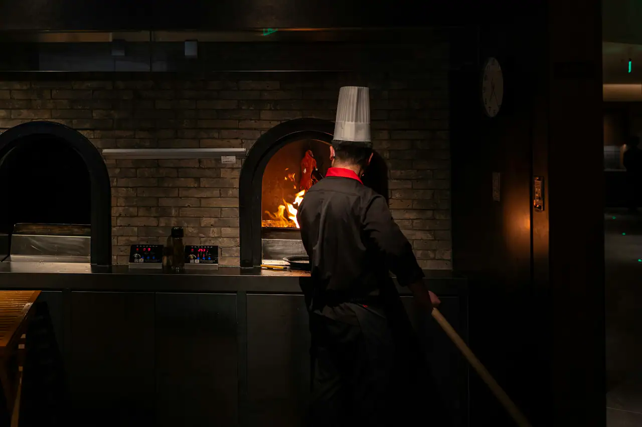 A chef in a tall white hat and black jacket, seen from behind, tending flames in an arched oven set into a dark brick wall