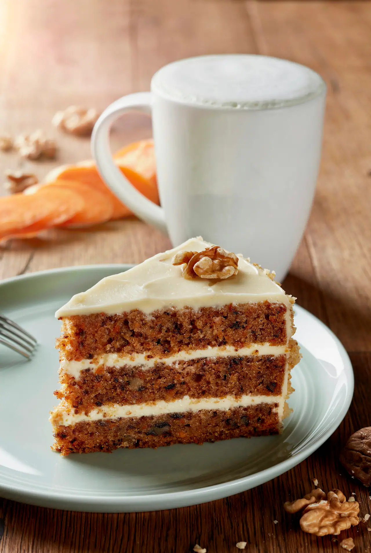 A three-layer carrot cake slice with cream cheese frosting and a walnut on a pale plate, beside a white mug of frothy coffee on a wooden table