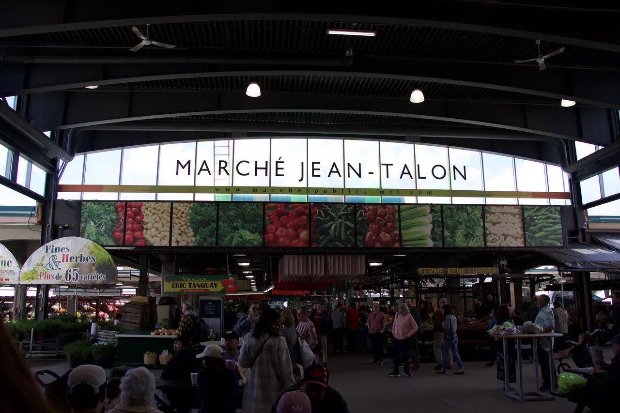 Inside the covered Marché Jean-Talon under the 'Marché Jean-Talon' sign, with vegetable murals above the vendor stalls and shoppers browsing below