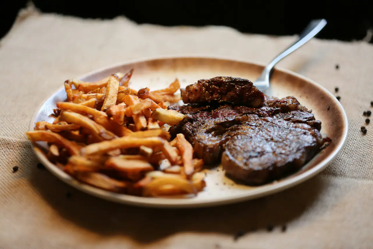 Sliced seared steak beside a pile of hand-cut fries on a rustic plate over burlap, with a fork and scattered peppercorns