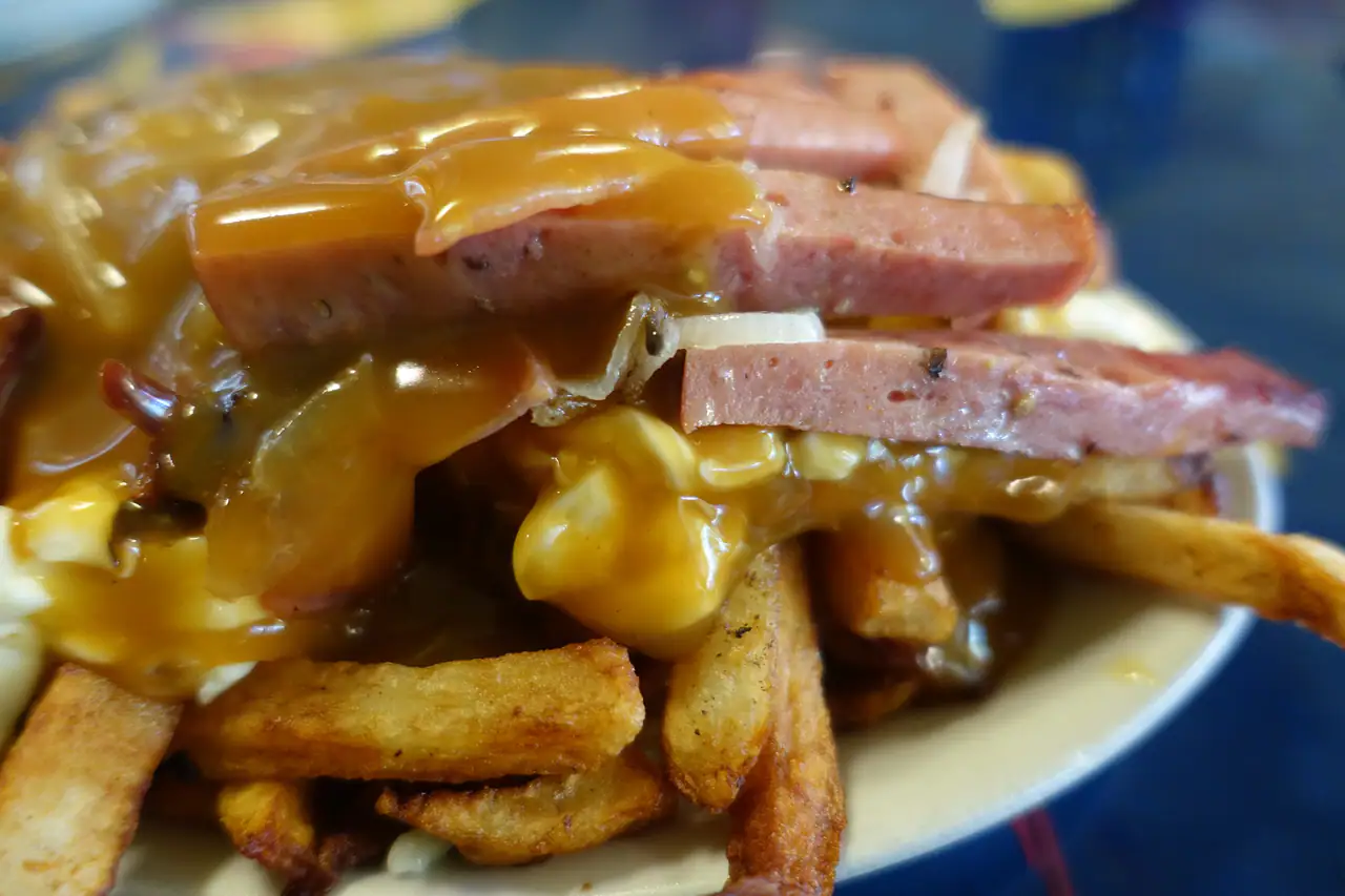 Close-up of a poutine at La Banquise: thick-cut fries, cheese curds and sliced sausage under glossy brown gravy on a cream plate