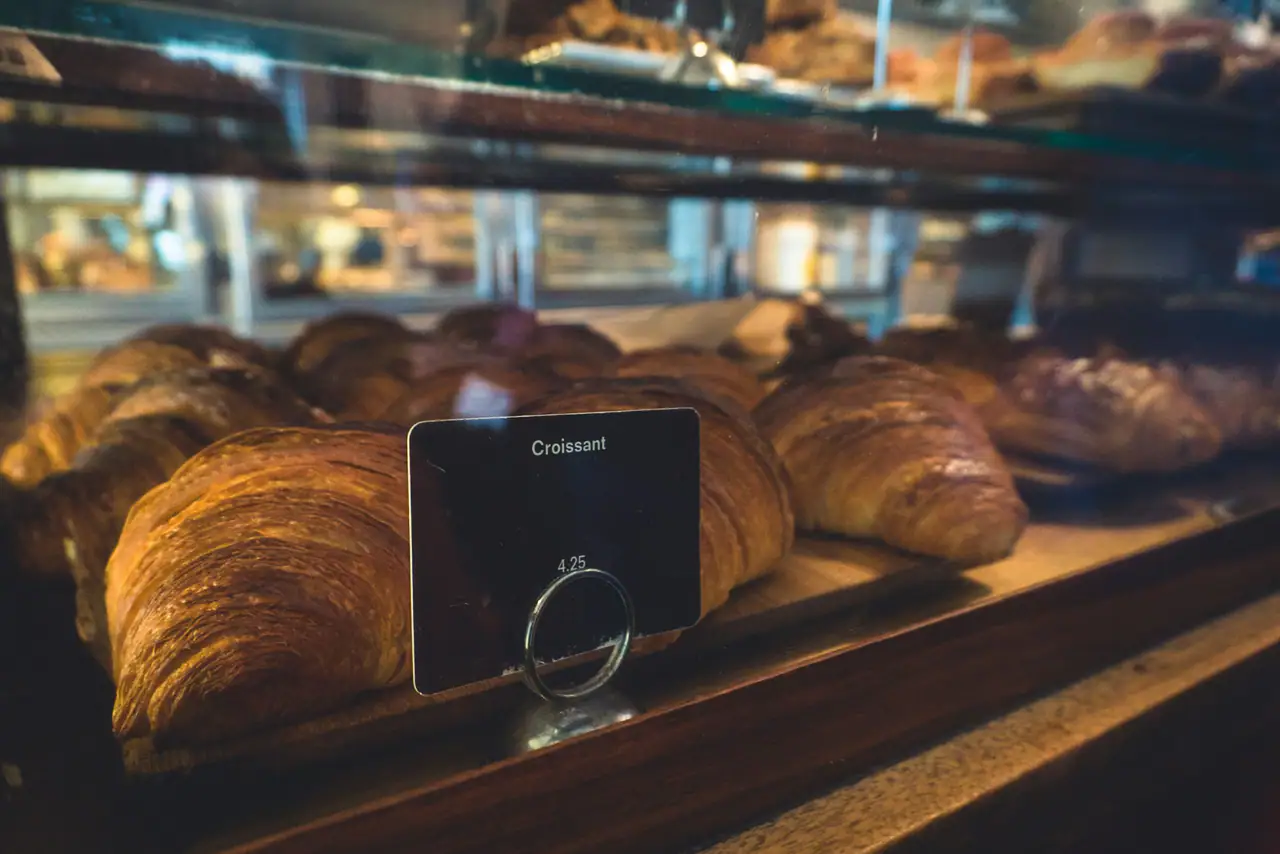 Golden croissants lined up on a wooden shelf behind a glass bakery display case, with a small black price card reading Croissant