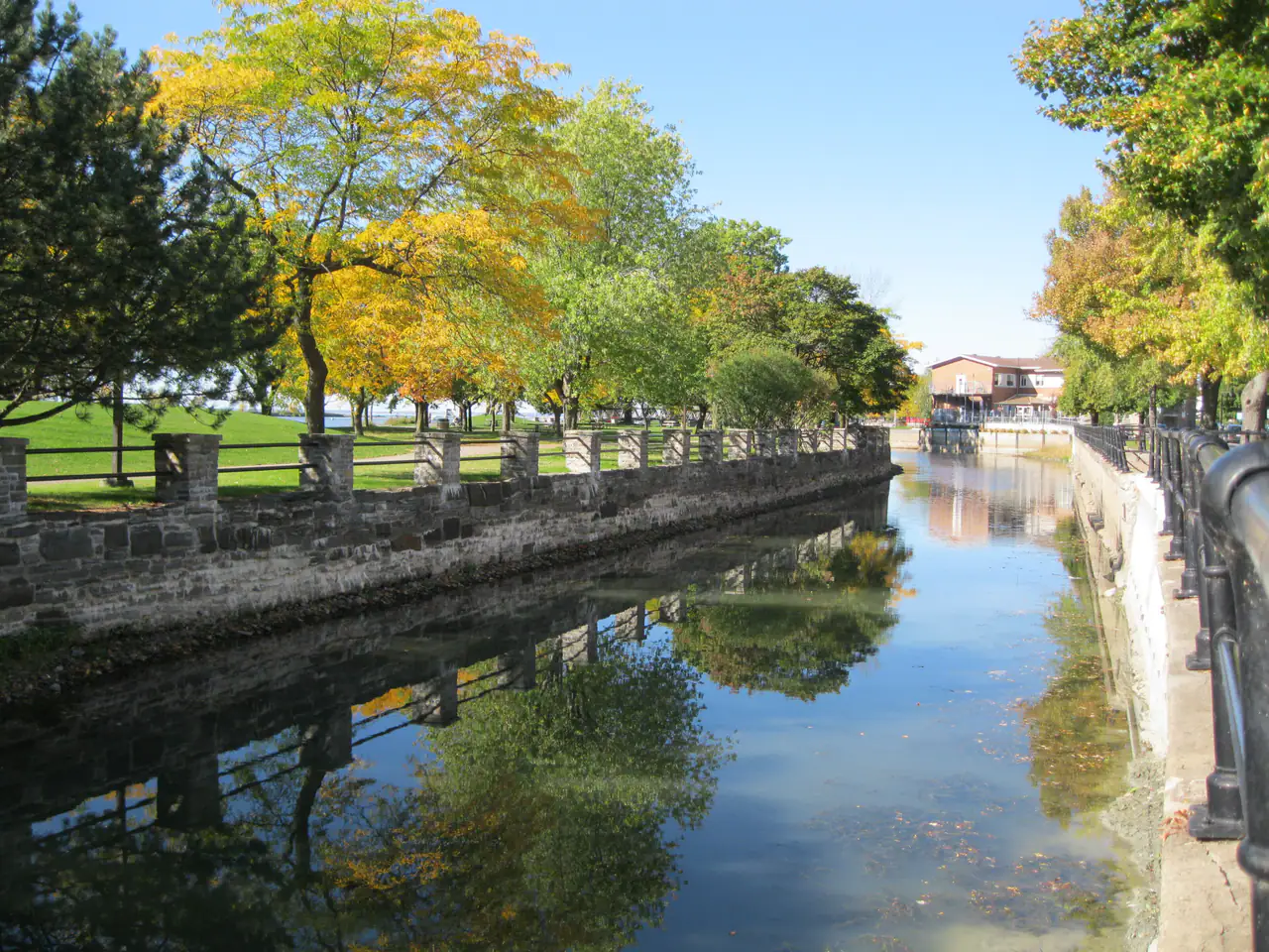 Autumn trees with yellow and green leaves line a stone-walled stretch of the Lachine Canal, reflected in still water under a blue sky