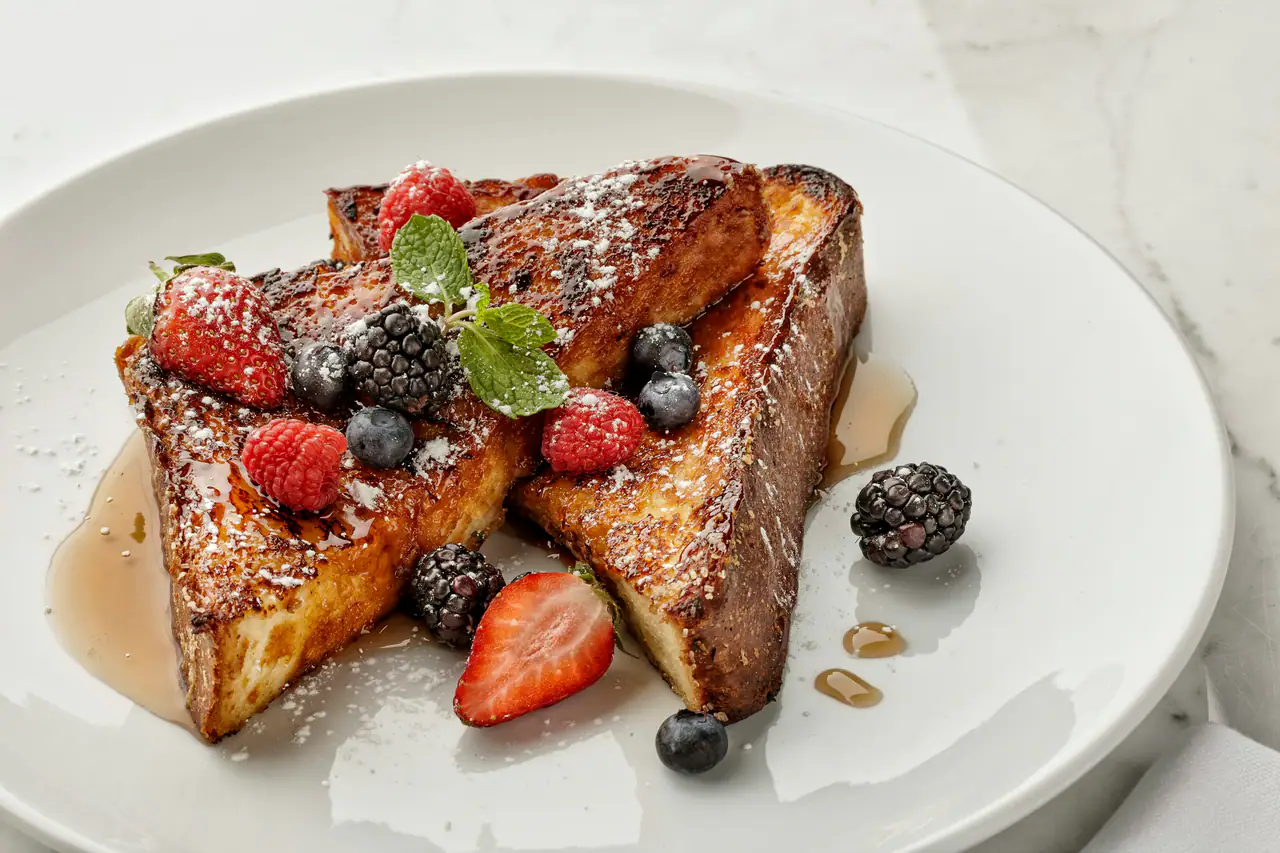 Thick slices of golden French toast dusted with powdered sugar, topped with raspberries, blackberries, blueberries and mint, with maple syrup pooled on a white plate
