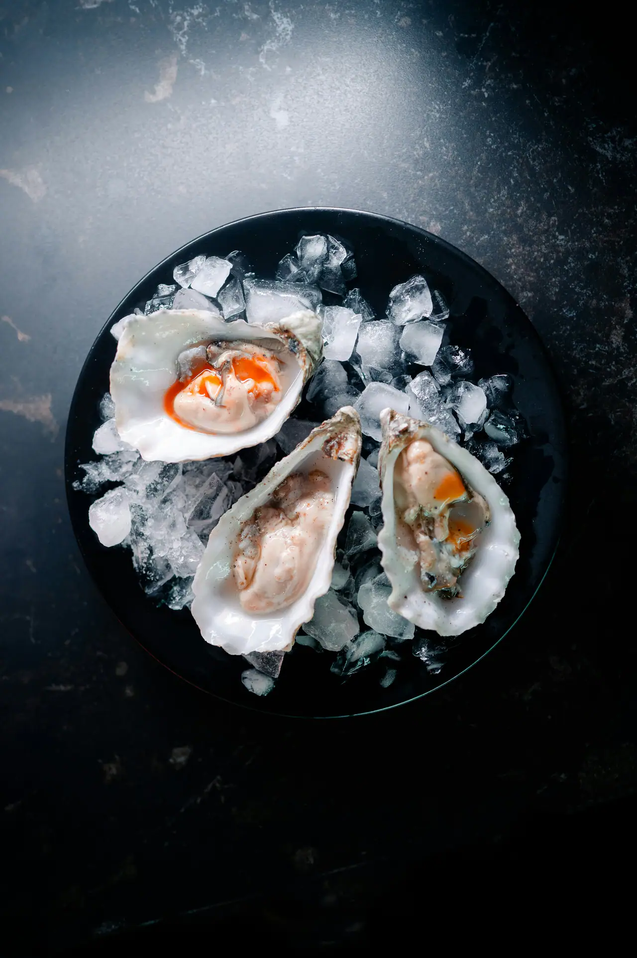 Three oysters on crushed ice on a black plate, set on a dark stone surface, from above