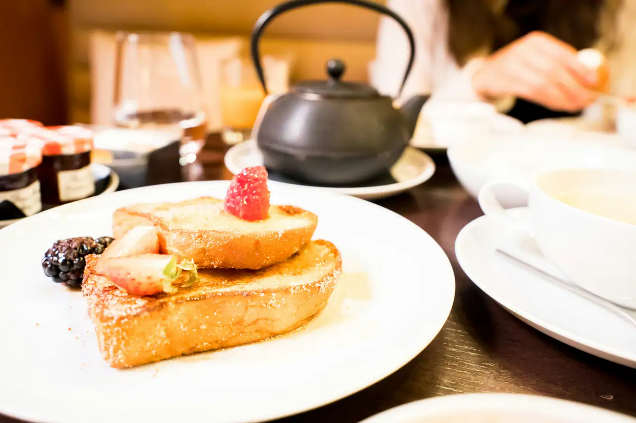 Thick slices of golden French toast topped with a raspberry, strawberry and blackberry on a white plate, with an iron teapot and a cup on a dark wooden table behind