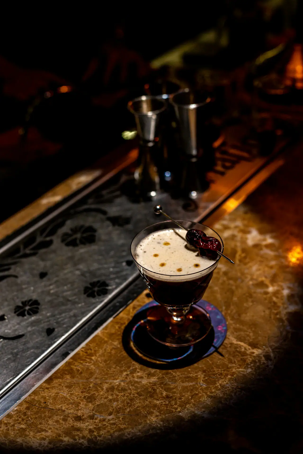 A frothy cocktail in a coupe glass garnished with a cherry on a skewer, set on a marble bar top in dim, amber light